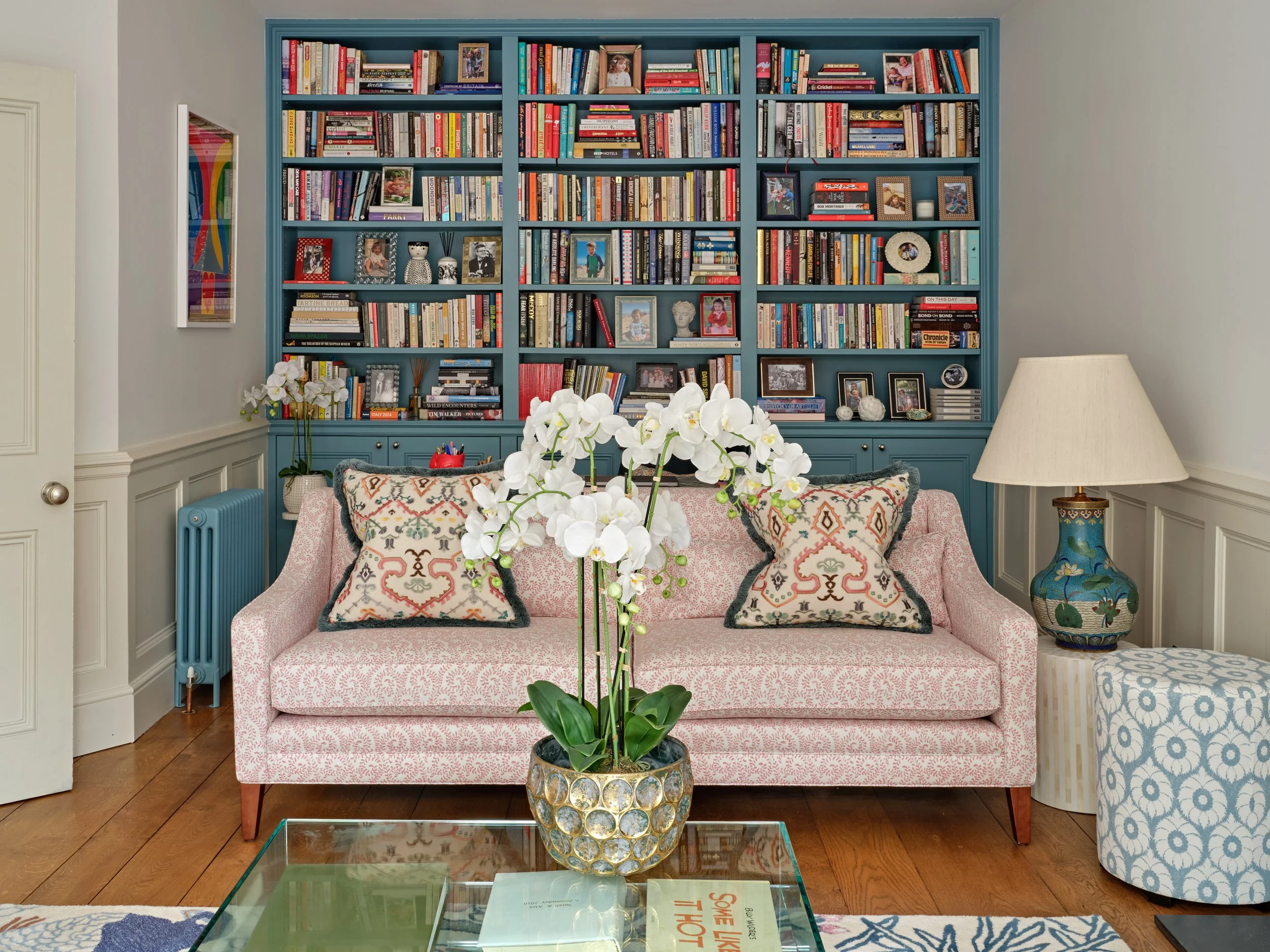 Living room with a blue bookshelf filled with books and photos, a pink patterned sofa with pillows, a glass coffee table with magazines, a vase with white orchids, and a lamp with a decorative base.