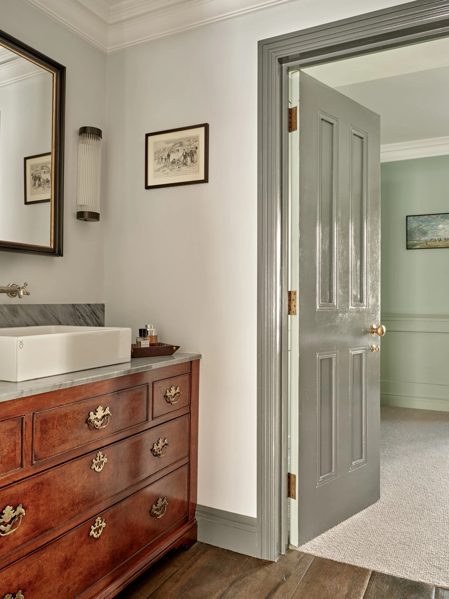 A bathroom vanity with wooden drawers and a marble countertop, a white vessel sink, a soap dispenser, picture frames on the wall, a mirror, and a wall-mounted light fixture. An open gray door leads to a room with green walls and beige carpet.