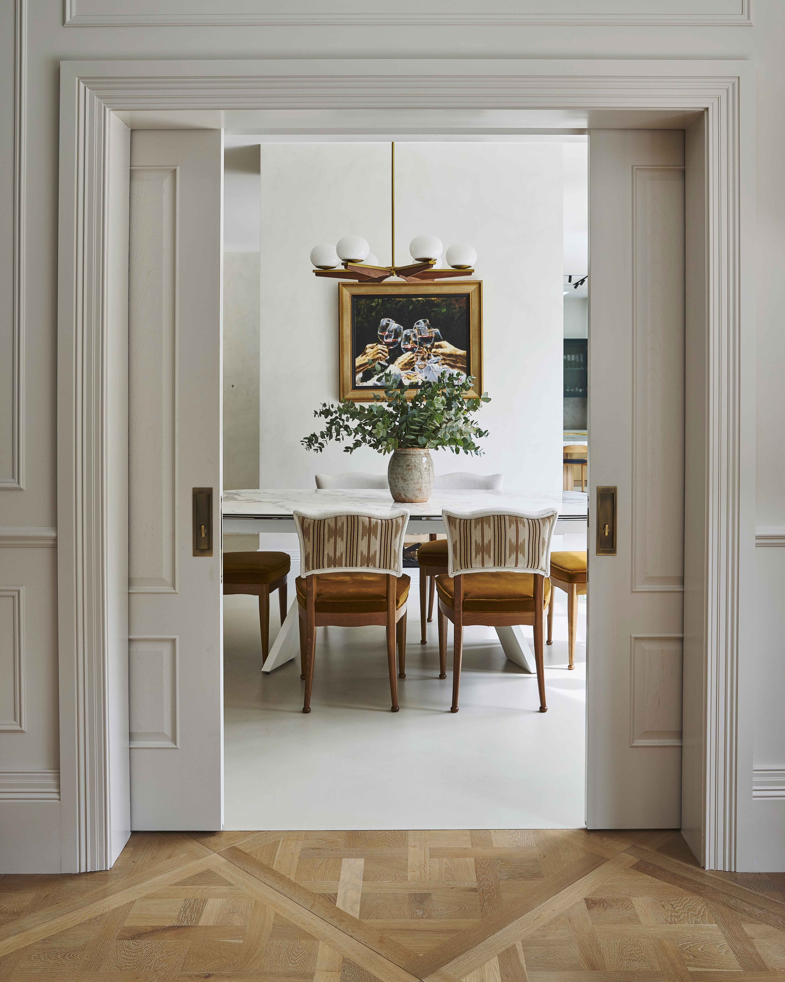 A dining room with a white marble table, four wooden chairs with striped upholstery, a large potted plant, a painting of people holding wine glasses, a modern chandelier, and is seen through an open doorway with detailed molding.