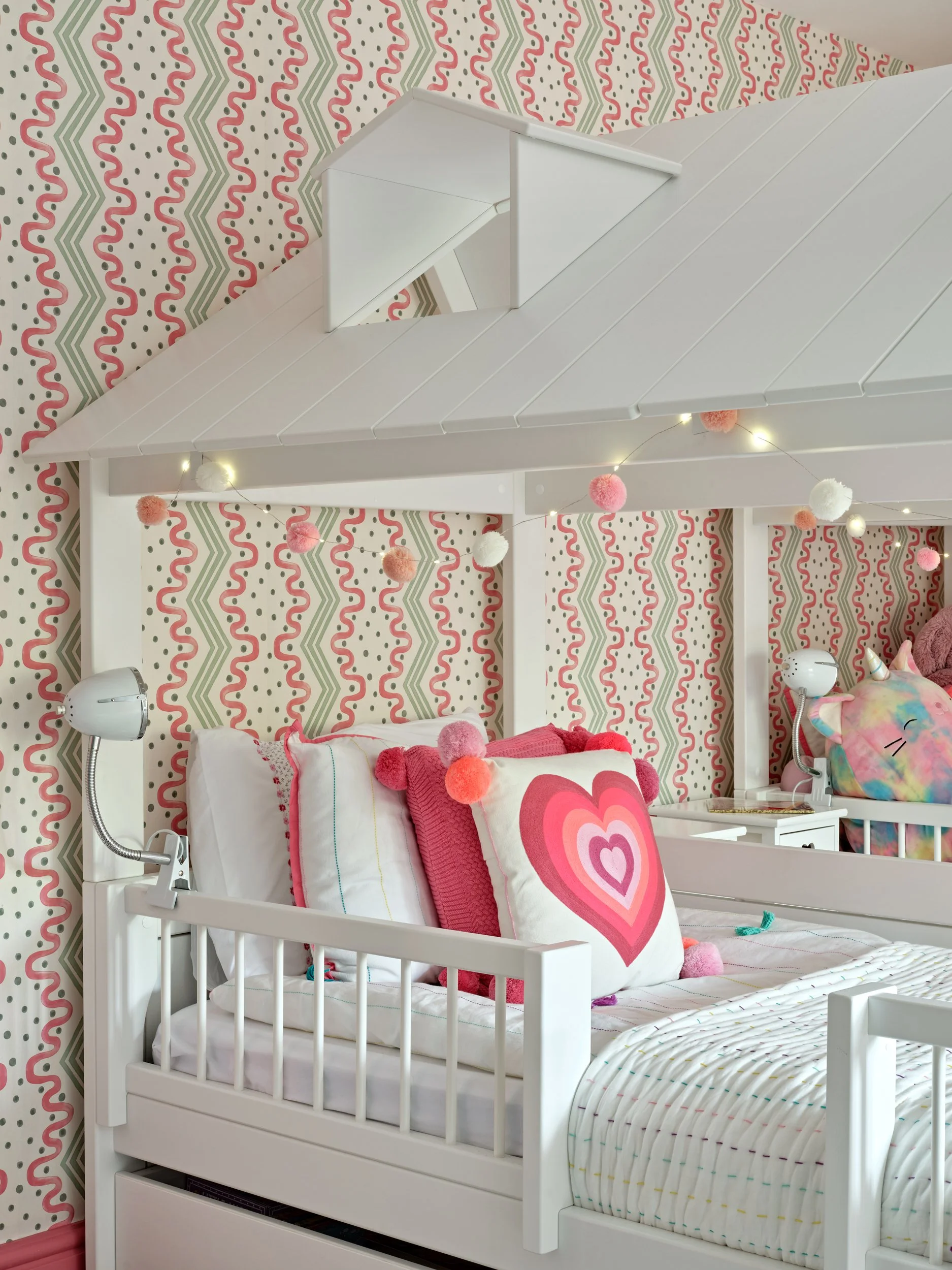 A child's bedroom with a white bed decorated with pillows featuring pink hearts, and a colorful plush unicorn toy. The background has a pink, red, green, and white patterned wallpaper, and a string of pink, white, and orange pom-pom lights hangs abov