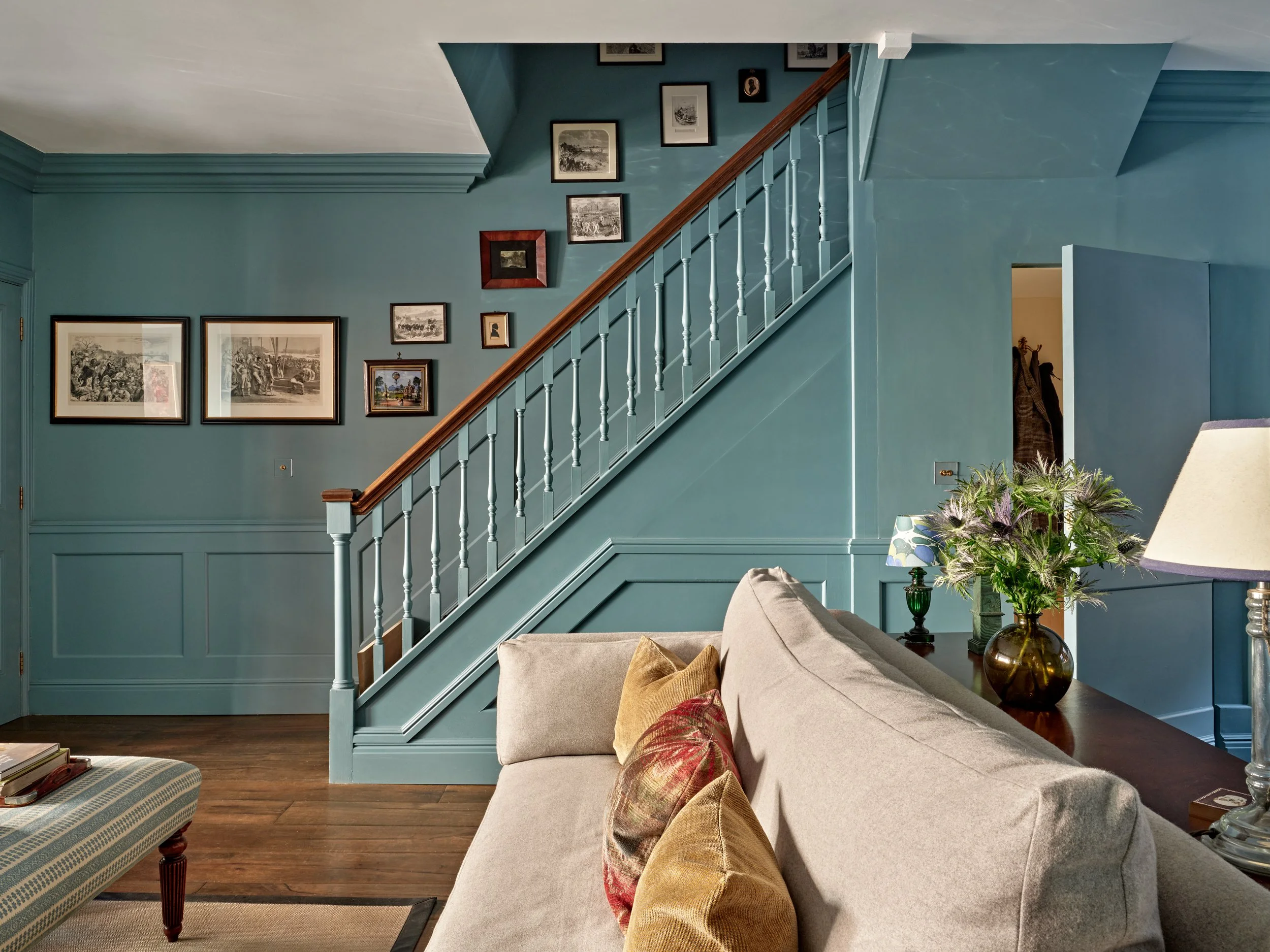 Living room with blue walls, decorated with framed pictures, a beige couch with colorful pillows, a glass table with a flower vase, and a staircase with white spindles and a wooden handrail.