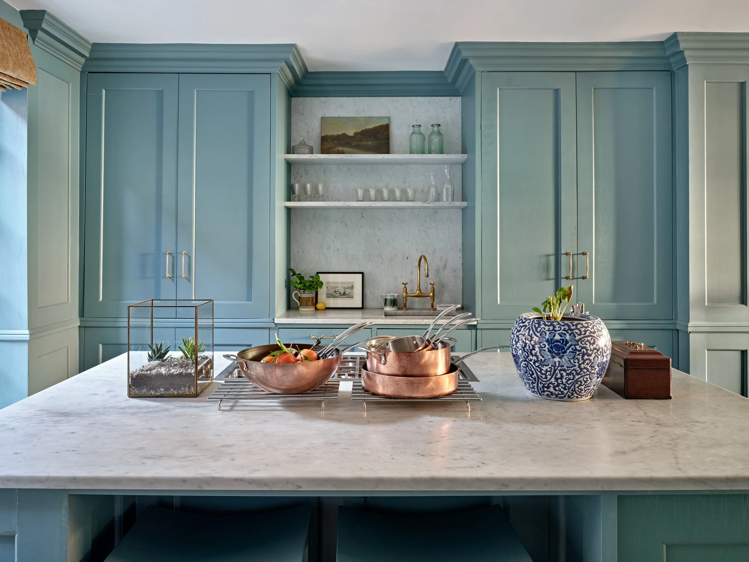 A kitchen with blue cabinetry, a marble countertop, open shelves with glass bottles and a landscape painting, and decorative items including a plant, framed picture, and a large blue and white ceramic vase.