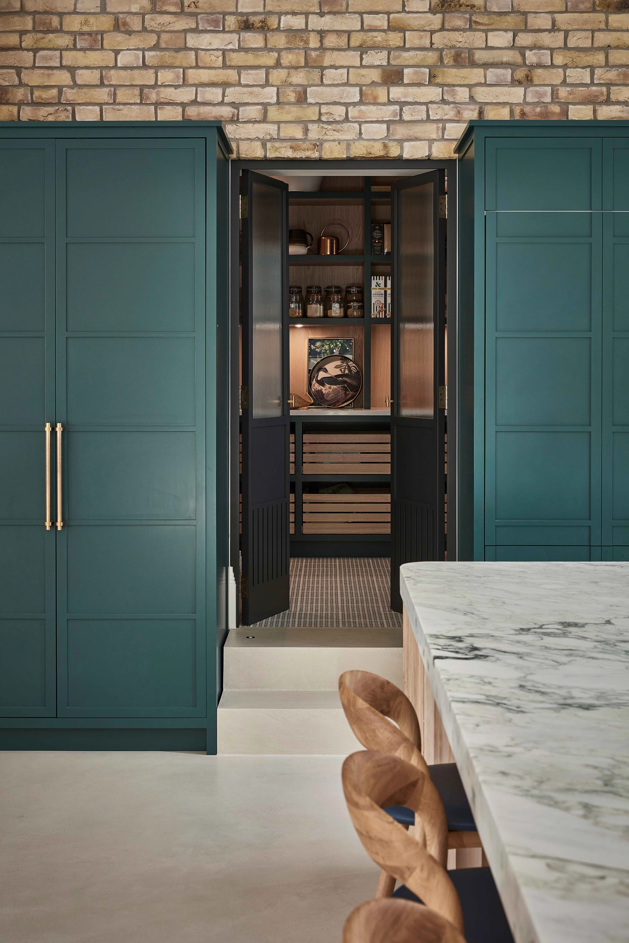 View of a modern kitchen with green cabinets, black door, marble table, wooden chairs, and a brick wall. An open pantry shows jars and kitchen items.