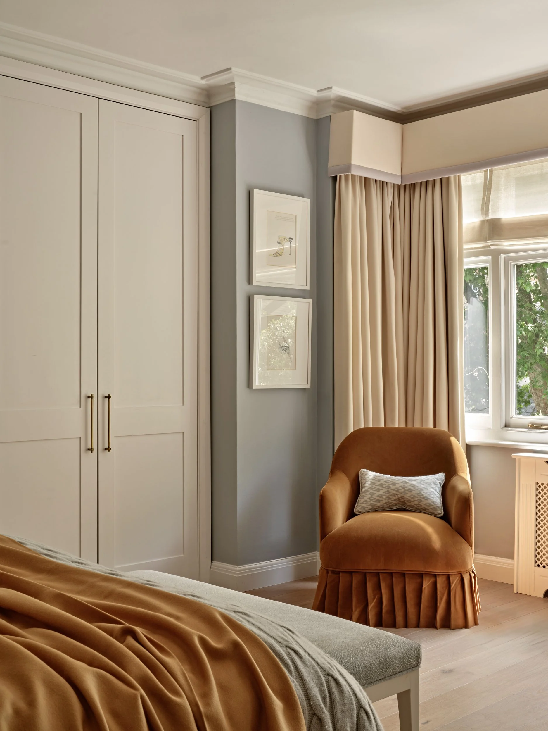 A cozy sitting area near a window with beige curtains, a brown armchair with a decorative pillow, and built-in white cabinets on a soft blue wall with two framed pictures.