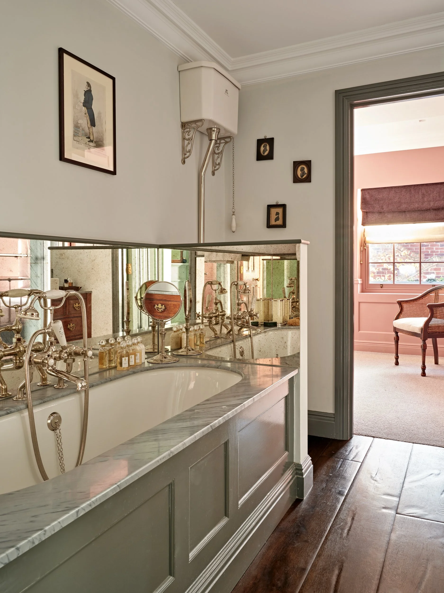 Vintage bathroom with a white bathtub, silver fixtures, small bottles, and a large mirror, with a doorway leading to a pink room with a carved wooden chair.