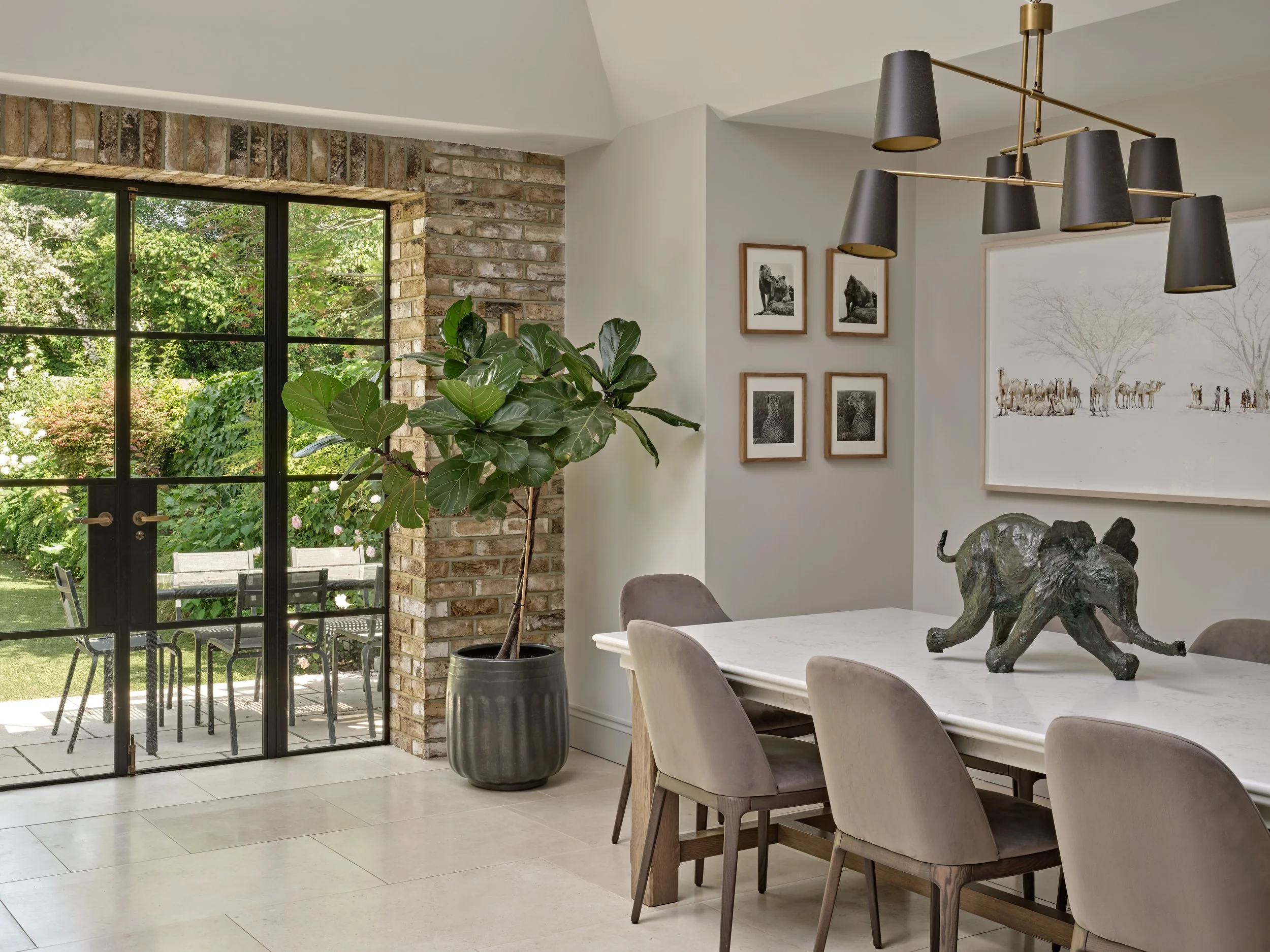 Interior view of a dining area with a white table, beige chairs, a large potted plant, black-framed glass door leading to an outdoor patio, art on the wall including black-and-white animal photographs and a large painting of wildlife, a black and bra