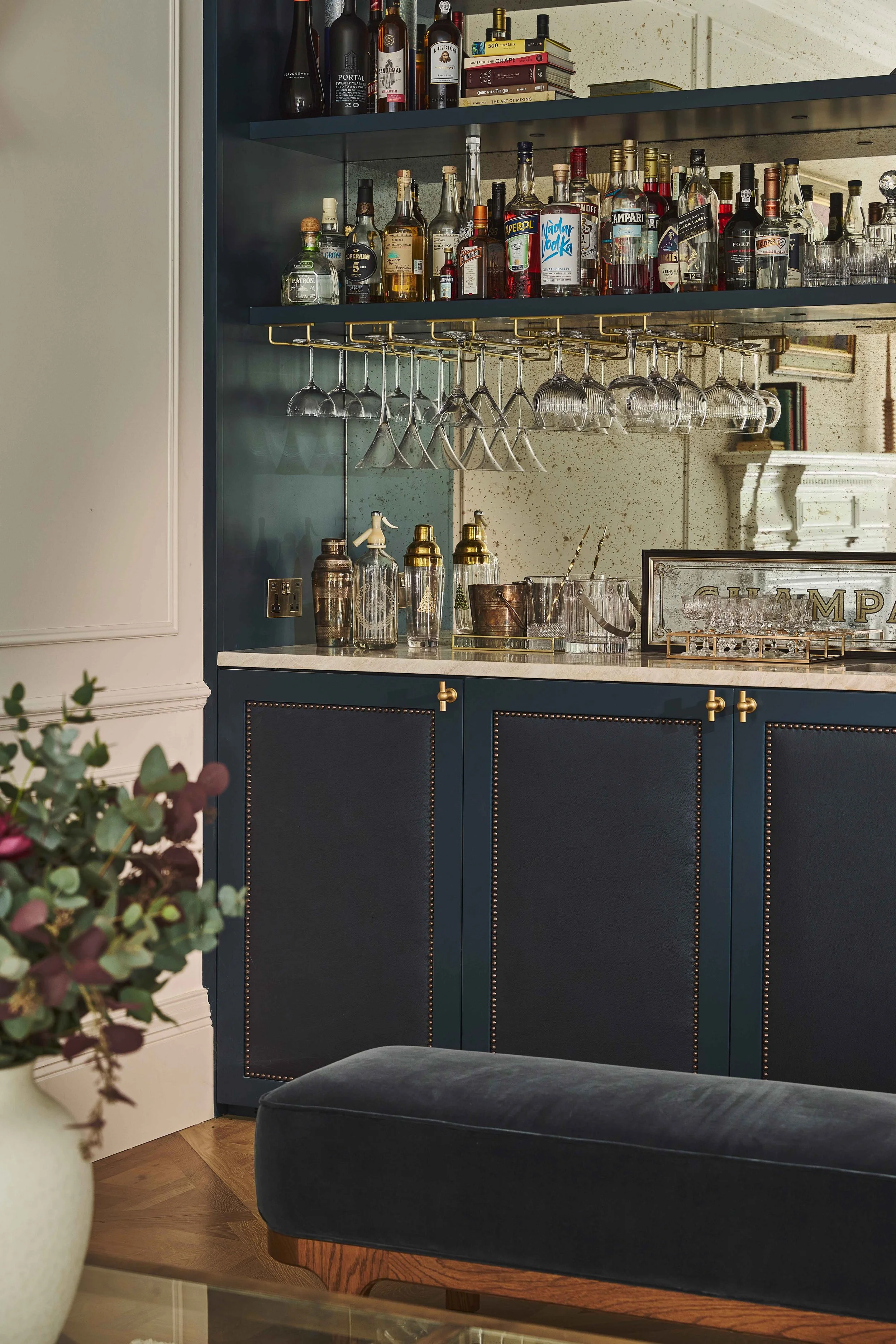 A kitchen or bar area with a dark blue cabinet that has two shelves filled with various bottles of alcohol and glasses hanging upside down. On top of the cabinet, there are stacked books and more bottles. A white countertop below the cabinet holds ad