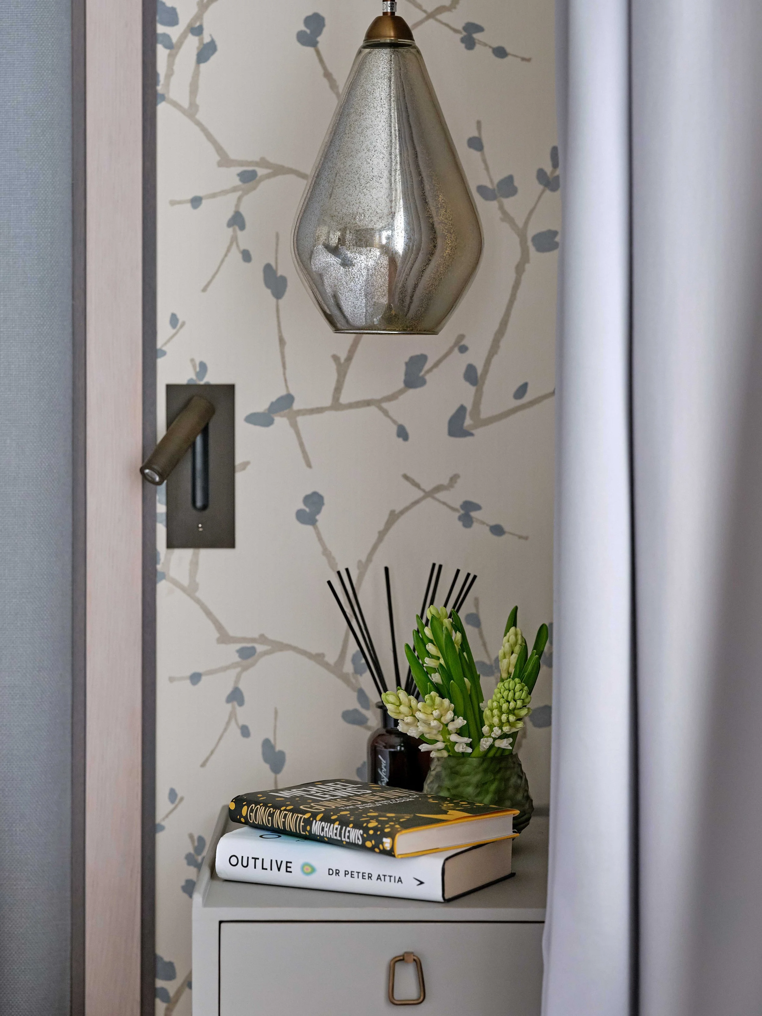 A white bedside table holding several books, a glass vase with fresh white flowers, and a decorative black reed diffuser. A diamond-shaped glass pendant light hangs above, and a patterned wallpaper with gray branches and leaves is in the background.