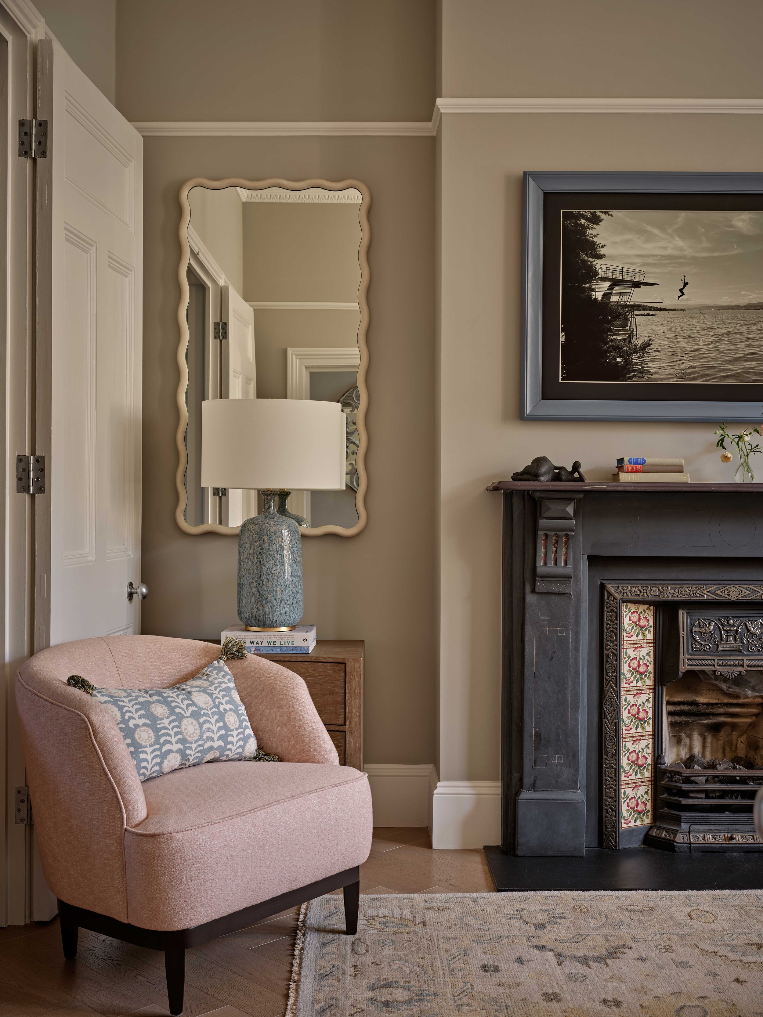 A cozy living room corner with a pink upholstered armchair with dark wooden legs, a light-patterned throw pillow, a side table with a textured blue ceramic table lamp and a book titled "The Way We Live," a large mirror with a wavy cream-colored frame, a framed black-and-white photograph of a seaside scene with a diving board and a swimmer, a dark fireplace mantel with decorative tiles, a small sculpture, a few books, a glass vase with flowers, and a patterned rug on dark hardwood flooring.