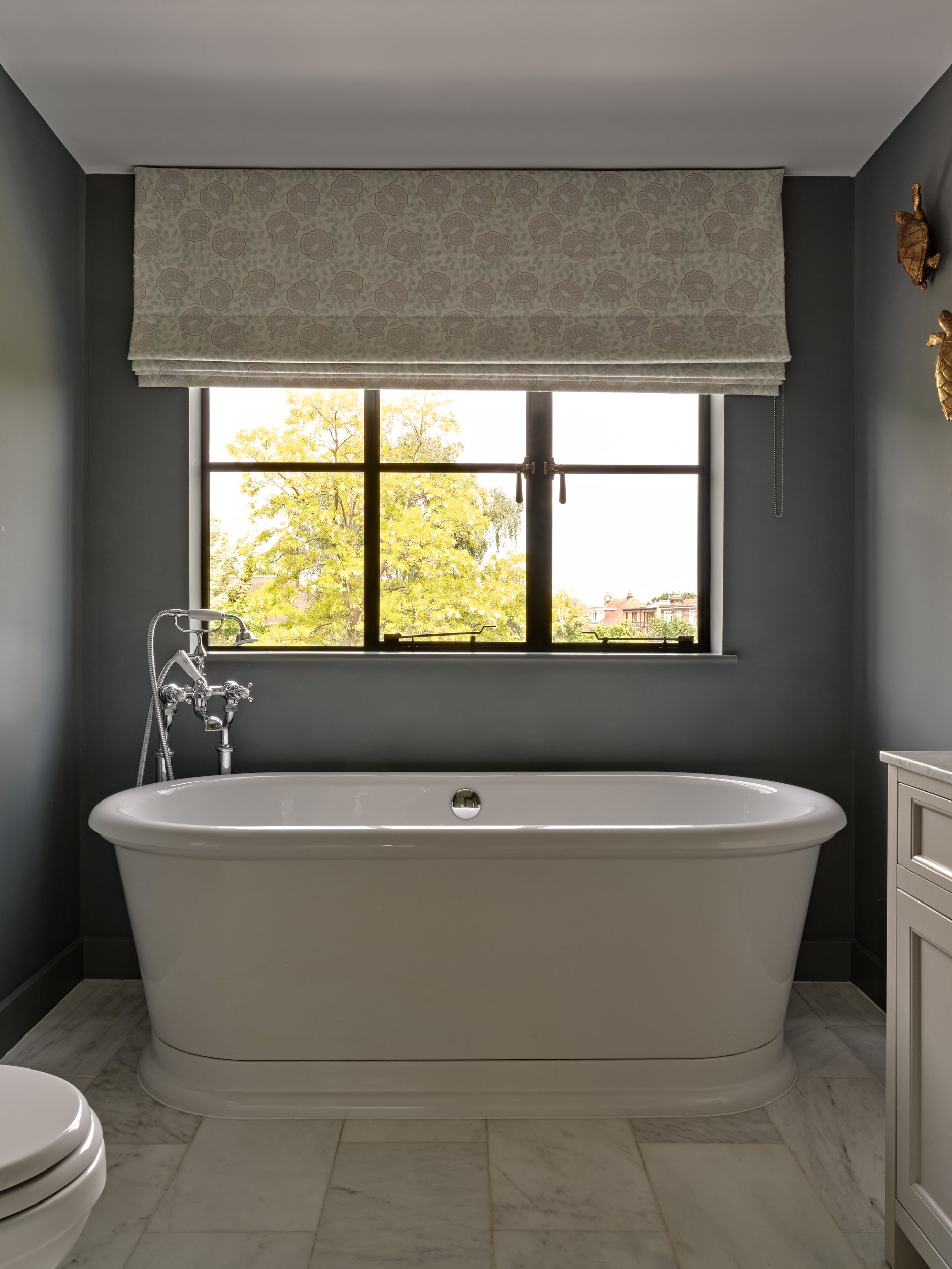 Bathroom with a white clawfoot bathtub, gray walls, tiled floor, window with patterned Roman shade, and a small cabinet.