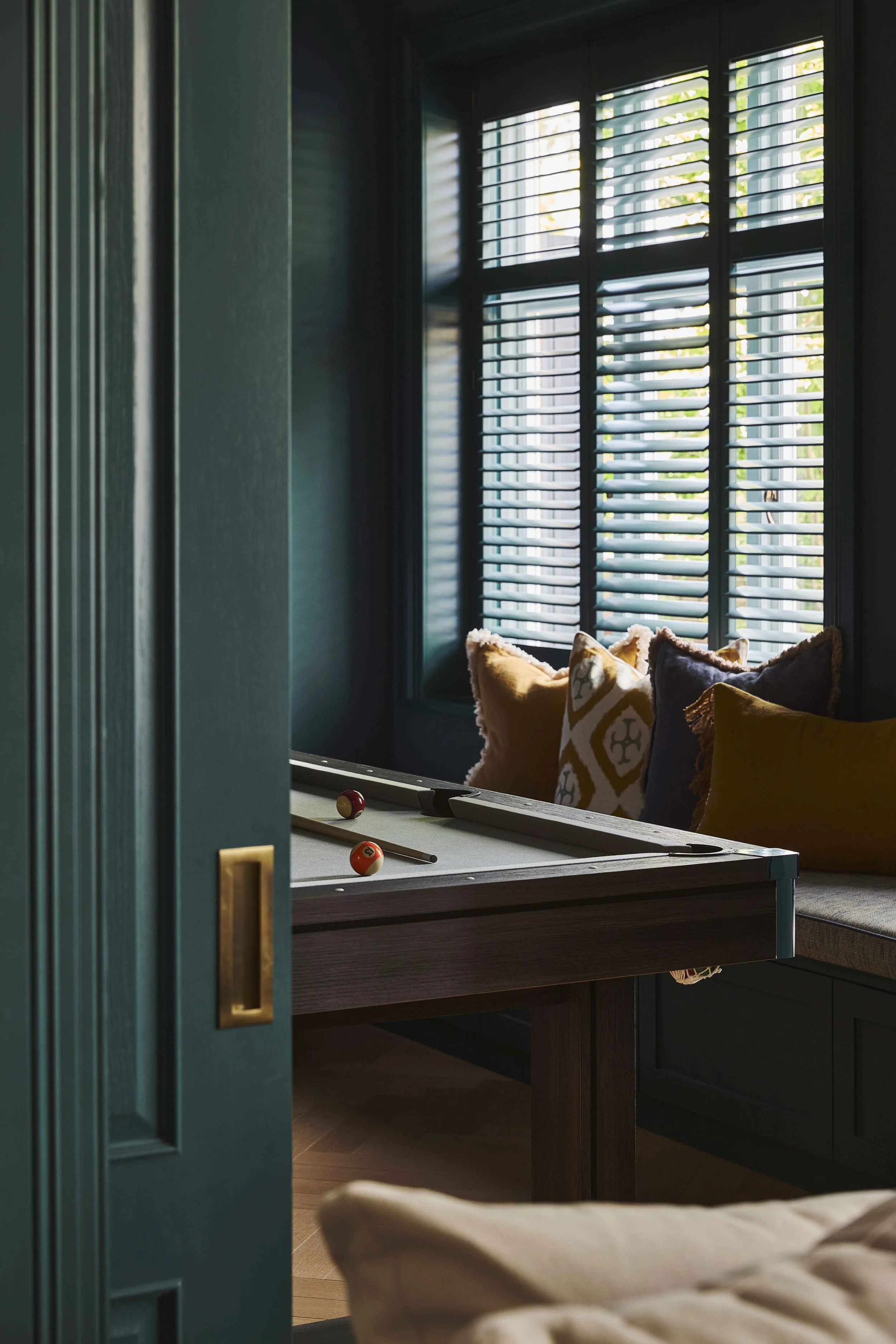 A view into a room featuring a pool table with balls and cues, pillows on a sofa, and a window with shutters letting in natural light.