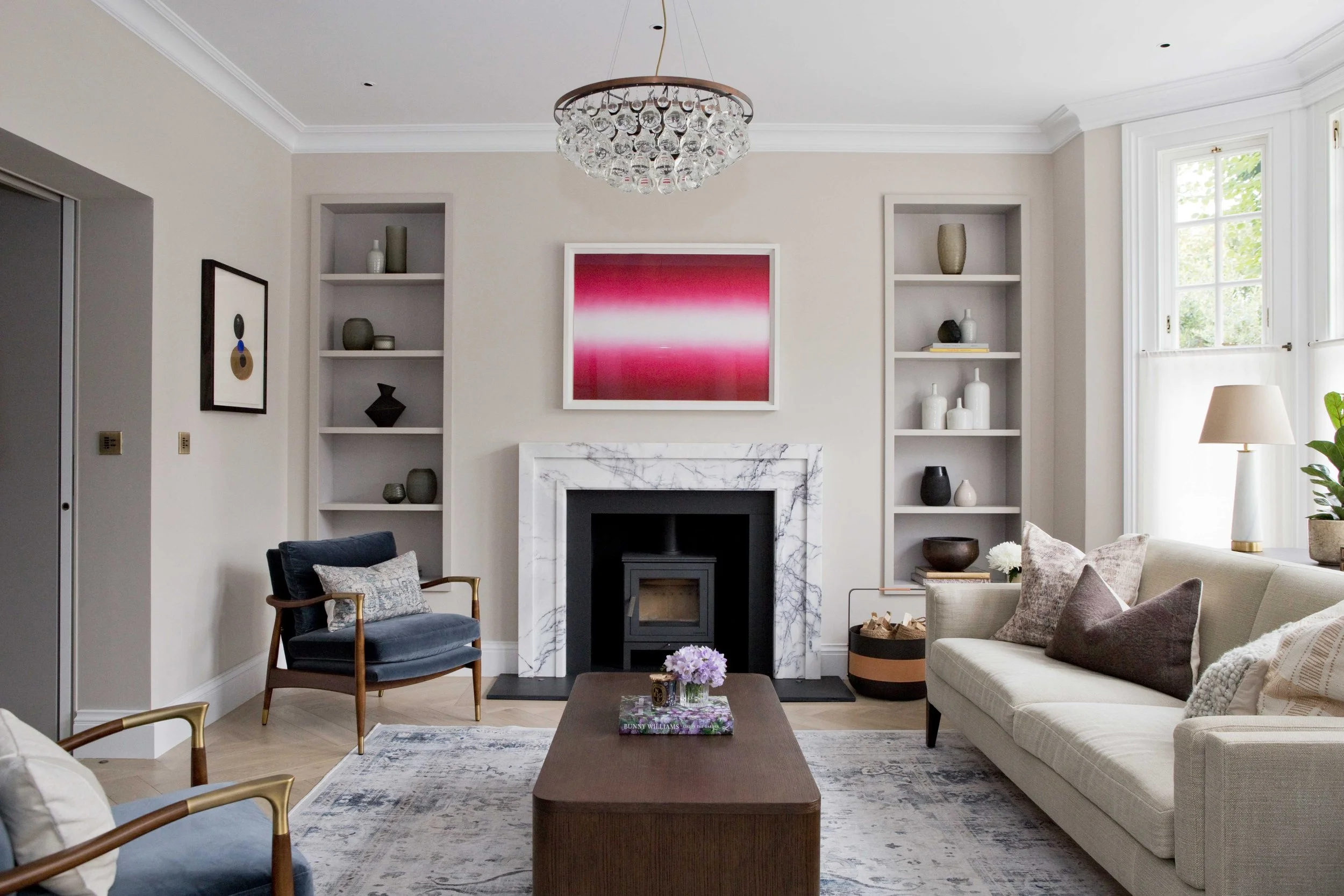 Living room with a fireplace framed in white marble, two built-in bookshelves with decorative items, a framed red abstract art piece above the fireplace, large windows with white trim, a white sofa with pillows, a dark blue armchair with a decorative