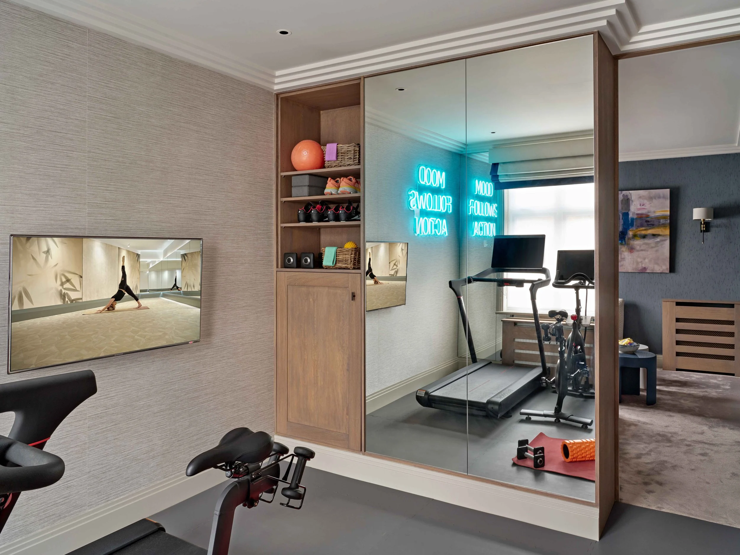 A modern home gym with exercise equipment including a treadmill, stationary bike, and exercise foam roller. The room has a large mirror, wooden shelving with workout supplies, and a neon sign on the wall.