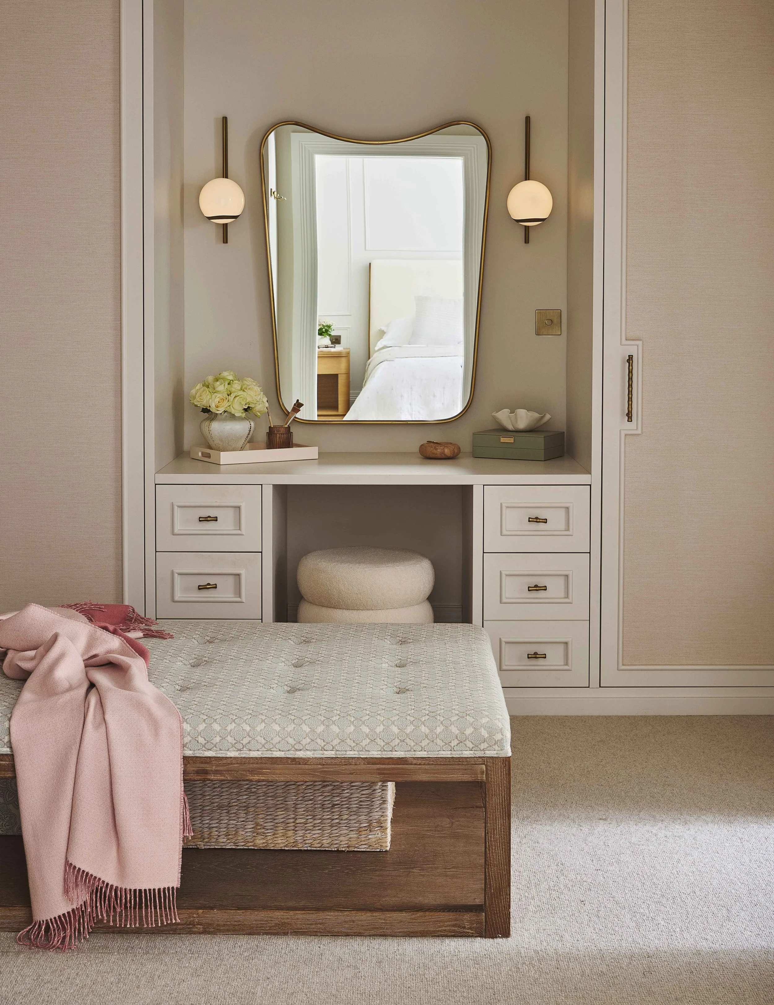 Bedroom decorative vanity area with a large mirror, two wall sconces, a vase of white flowers, a small bowl, a box, and two stacked stools, with a bed visible in the reflection.