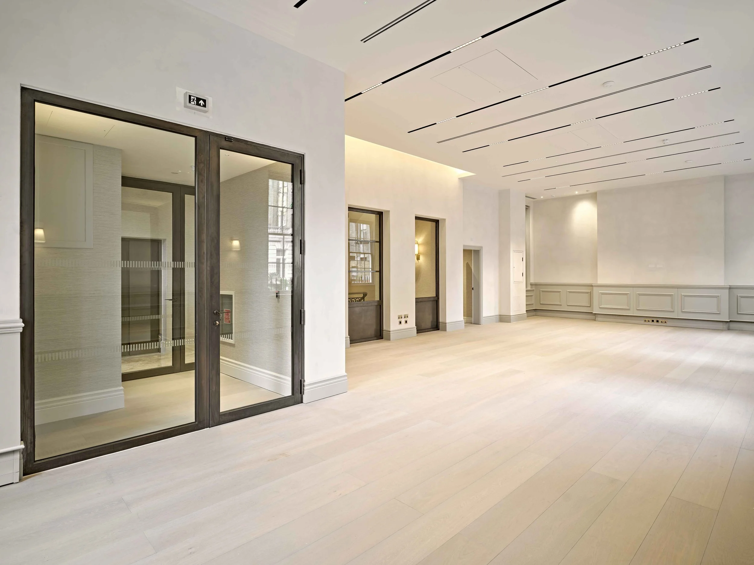 Modern interior space with white walls, light-colored wood flooring, and large windows. There are glass doors with dark frames, wall-mounted lights, and coffered ceiling with linear lighting.