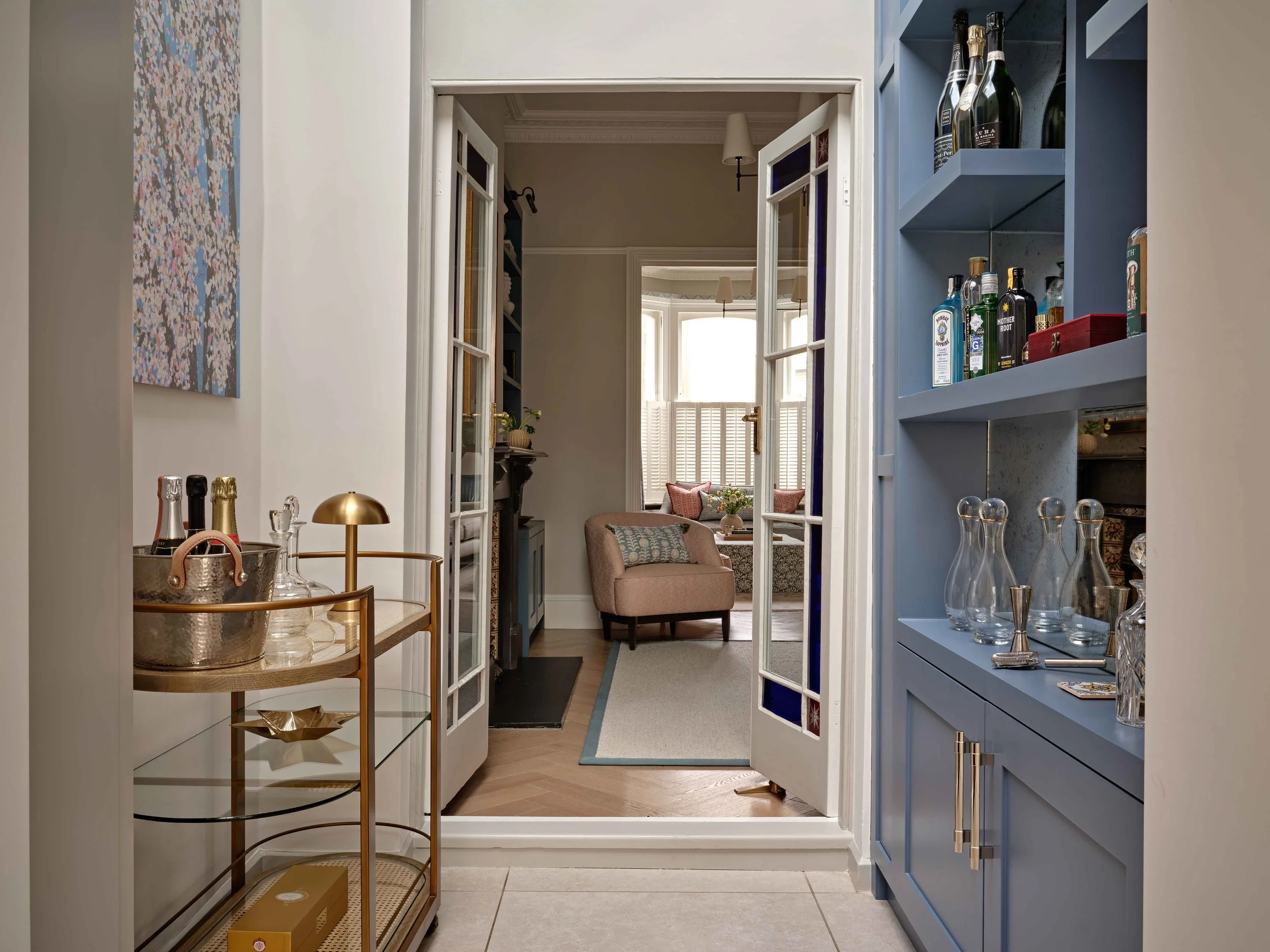 View from a kitchen opening into a living room, with a blue cabinet on the right displaying liquor bottles and glassware, a gold tray and a basket with champagne bottles on a cart on the left, and a cozy seating area with a beige armchair, pillows, a