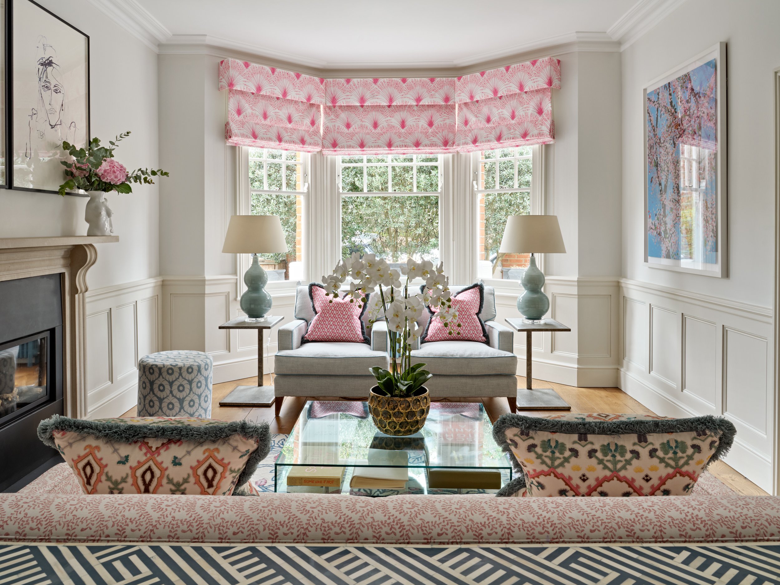 A living room with a large window with pink patterned Roman shades, white walls with crown molding, a gray sofa with pink and gray throw pillows, two matching table lamps on side tables, a glass coffee table with a decorative bowl, two upholstered ar