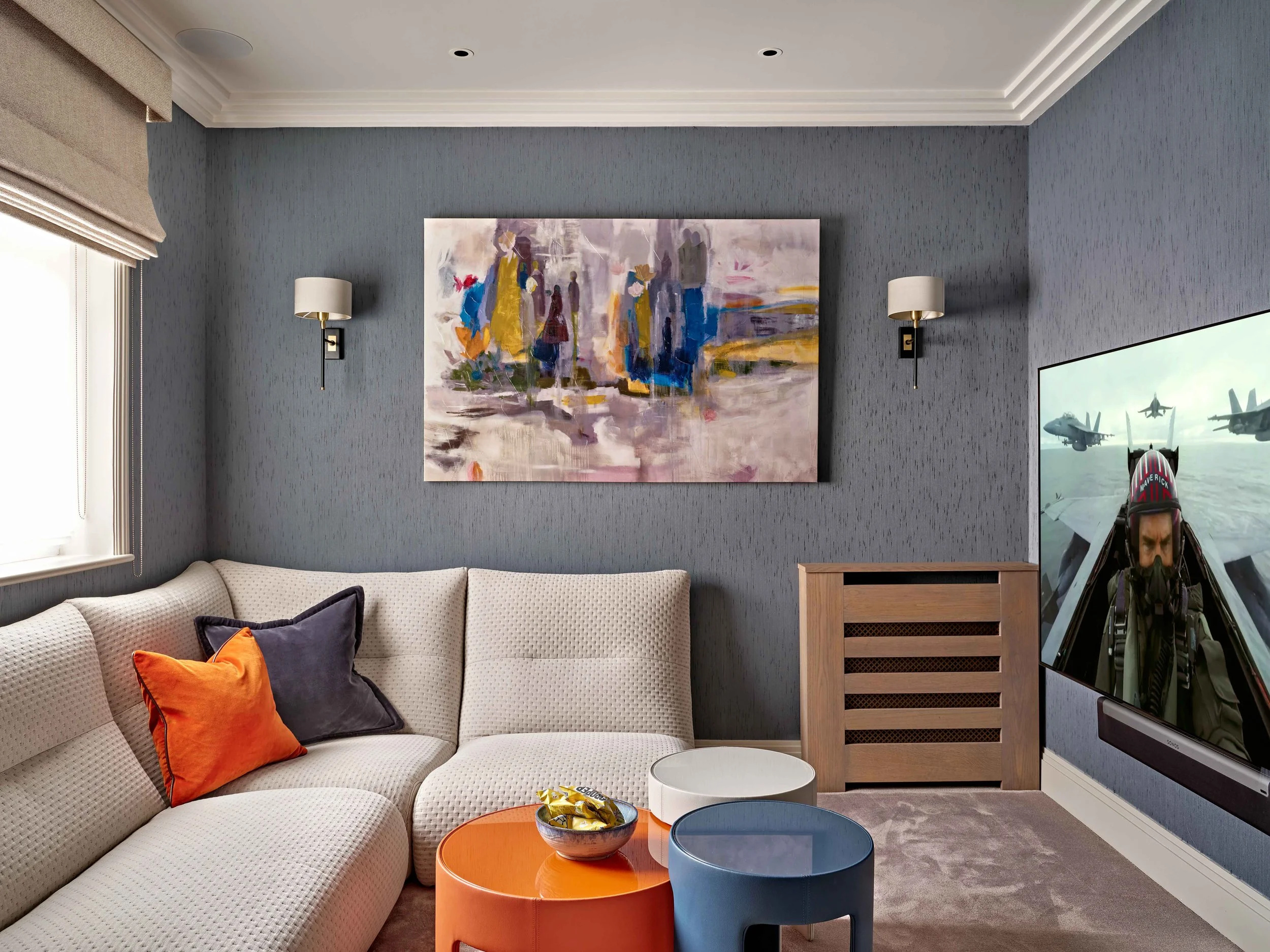 A living room with a beige sofa, colorful pillows, blue and orange side tables, a textured blue wall with a large abstract painting, wall sconces, a window with beige Roman shades, and a TV showing fighter jets.