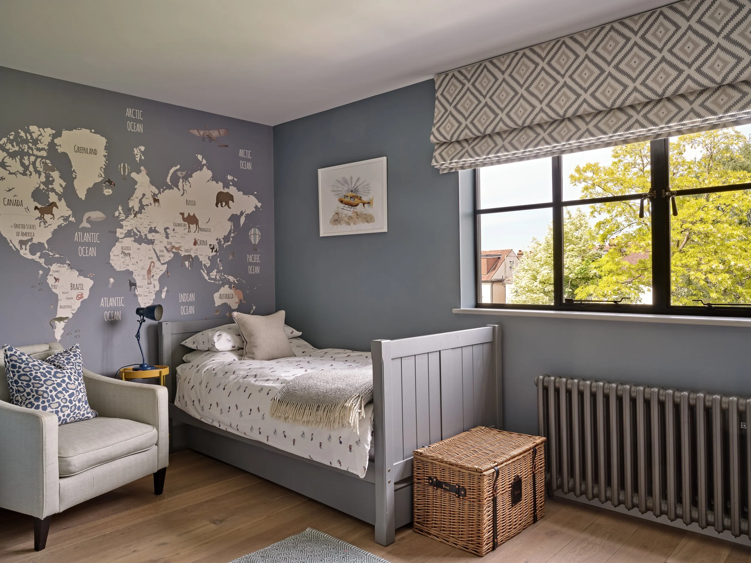 A bedroom with a white bed against a blue wall, decorated with a world map mural with animals and landscapes, and a window with patterned white curtains. There's a beige armchair with pillows, a wicker storage box, and a radiator.