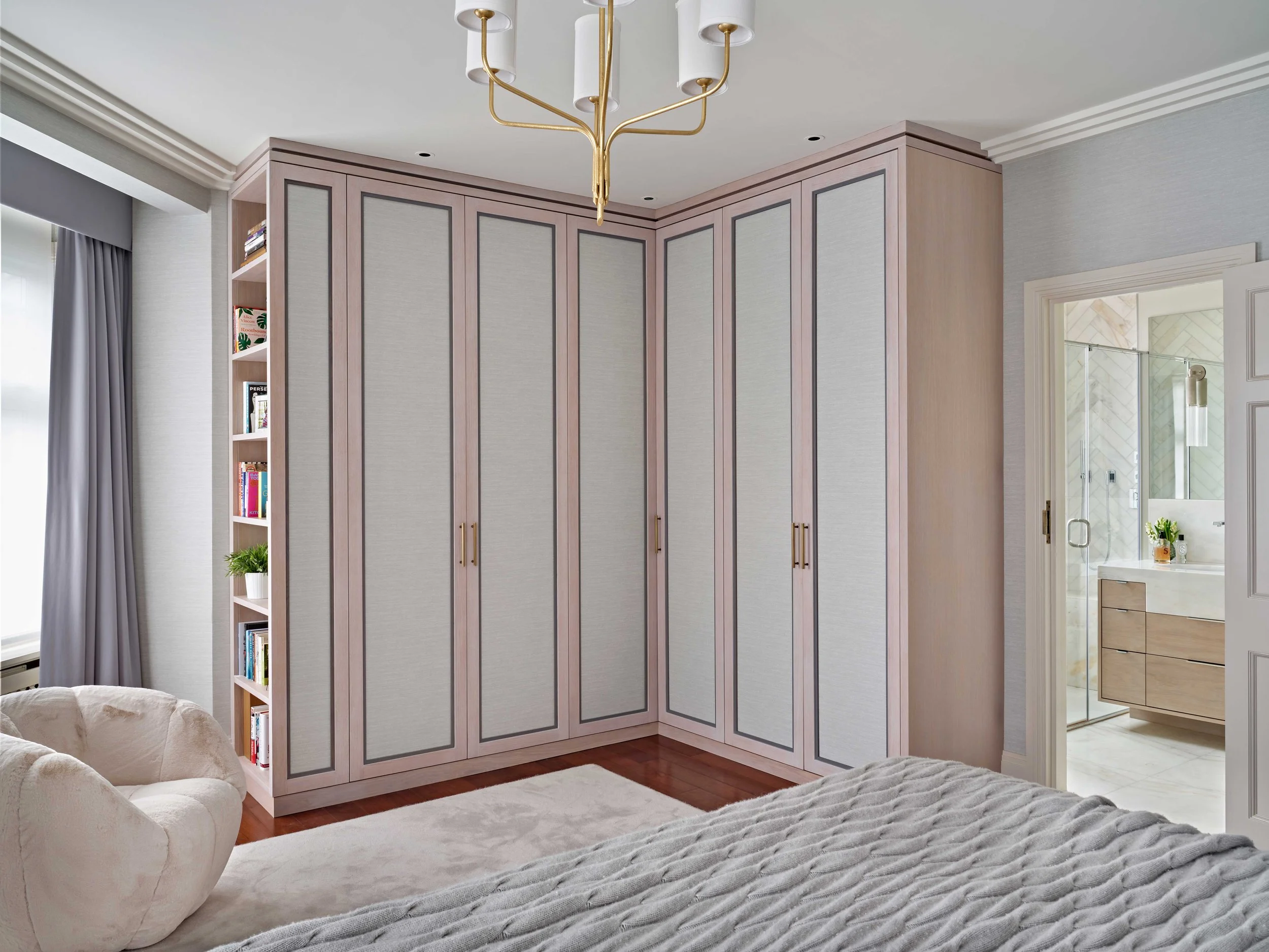 A bedroom with a tall built-in closet with gray panels and gold handles, a white ceiling with crown molding, a gold chandelier, and a window with gray curtains. There is a beige upholstered chair and a bed with a textured gray blanket visible in the 