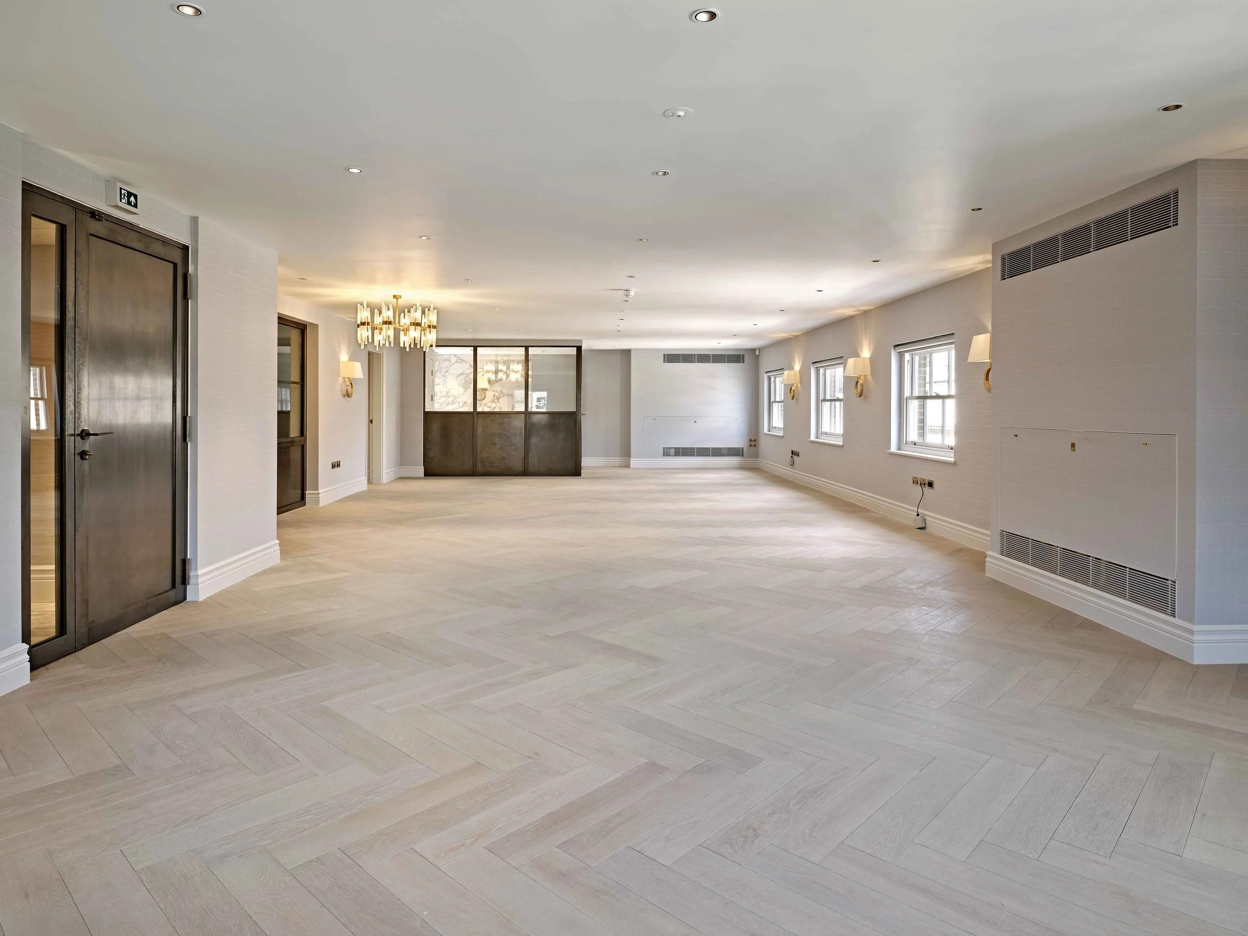 Empty spacious room with light wood flooring, white walls, multiple windows, wall-mounted lamps, a chandelier, and ventilation vents.