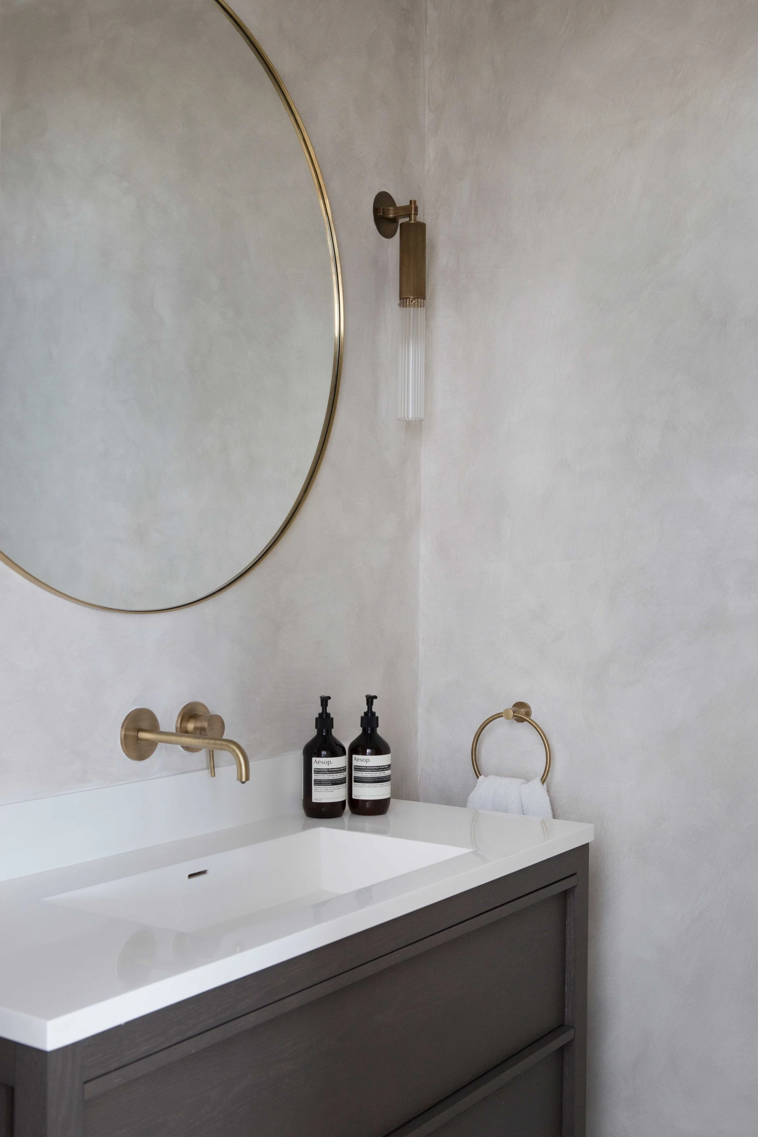 Modern bathroom vanity with a large round mirror, a wall-mounted gold faucet, black soap dispensers, a towel ring with a white towel, and gold wall-mounted showerhead and light fixture. The vanity has a white countertop and dark wood cabinetry.