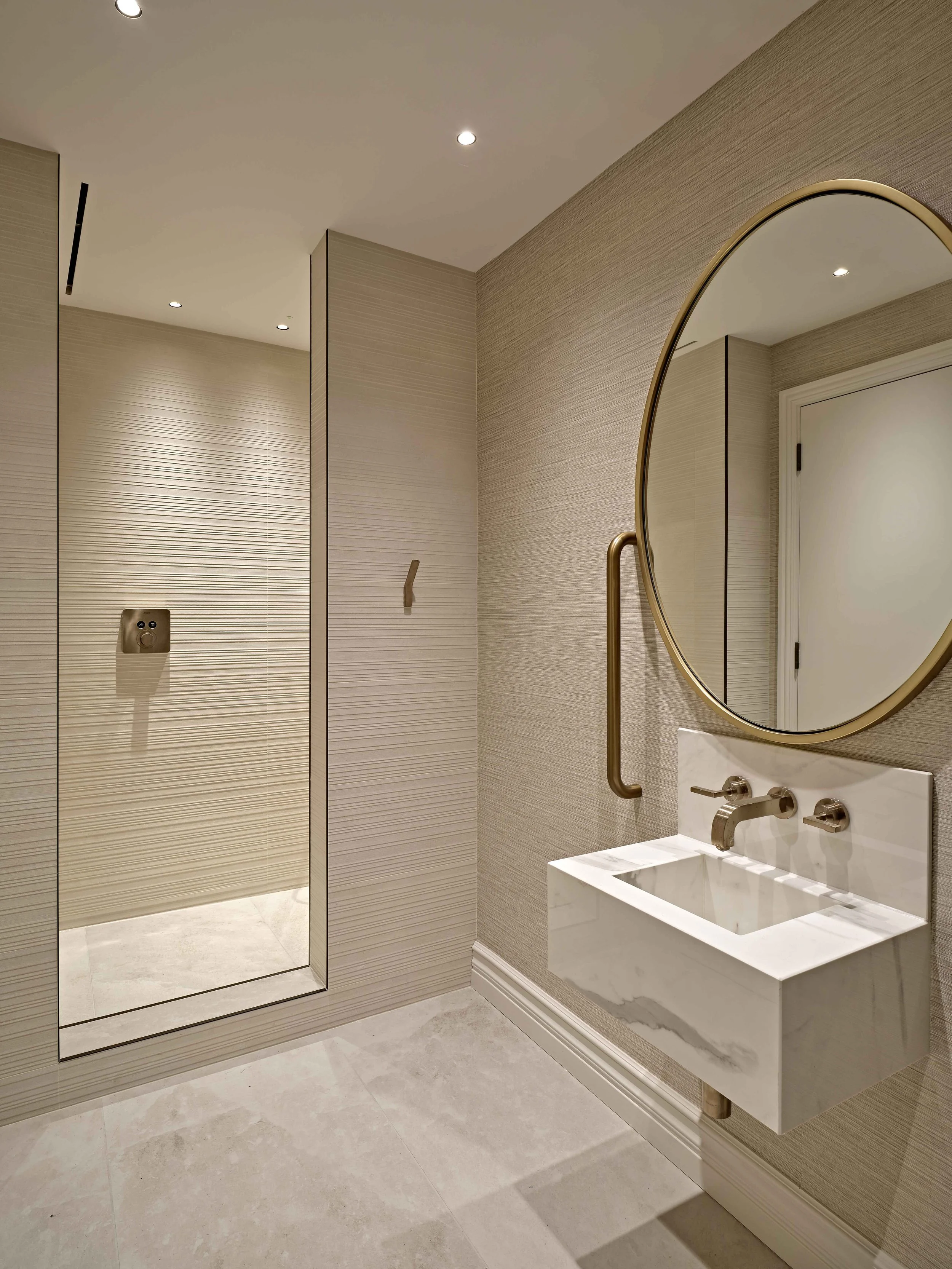 Modern bathroom with a large mirror, white marble sink, brass fixtures, textured beige wall, shower with glass door, and stone tile flooring.