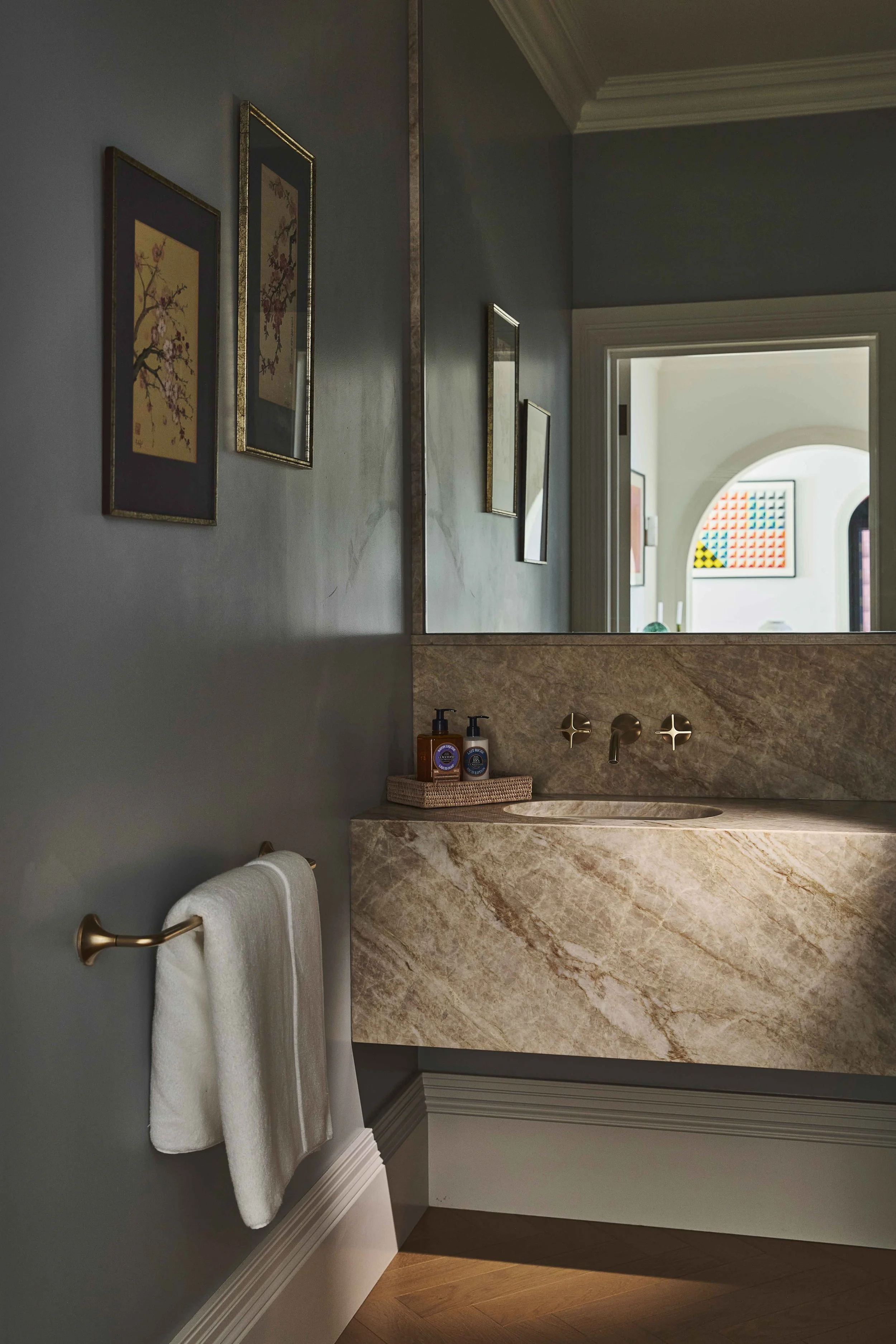 A bathroom with a large mirror above a marble vanity, containing two soap dispensers. The wall is painted a soft gray, and several framed art pieces hang on the wall. A towel hangs on a gold towel rod beside the vanity, and a doorway in the backgroun