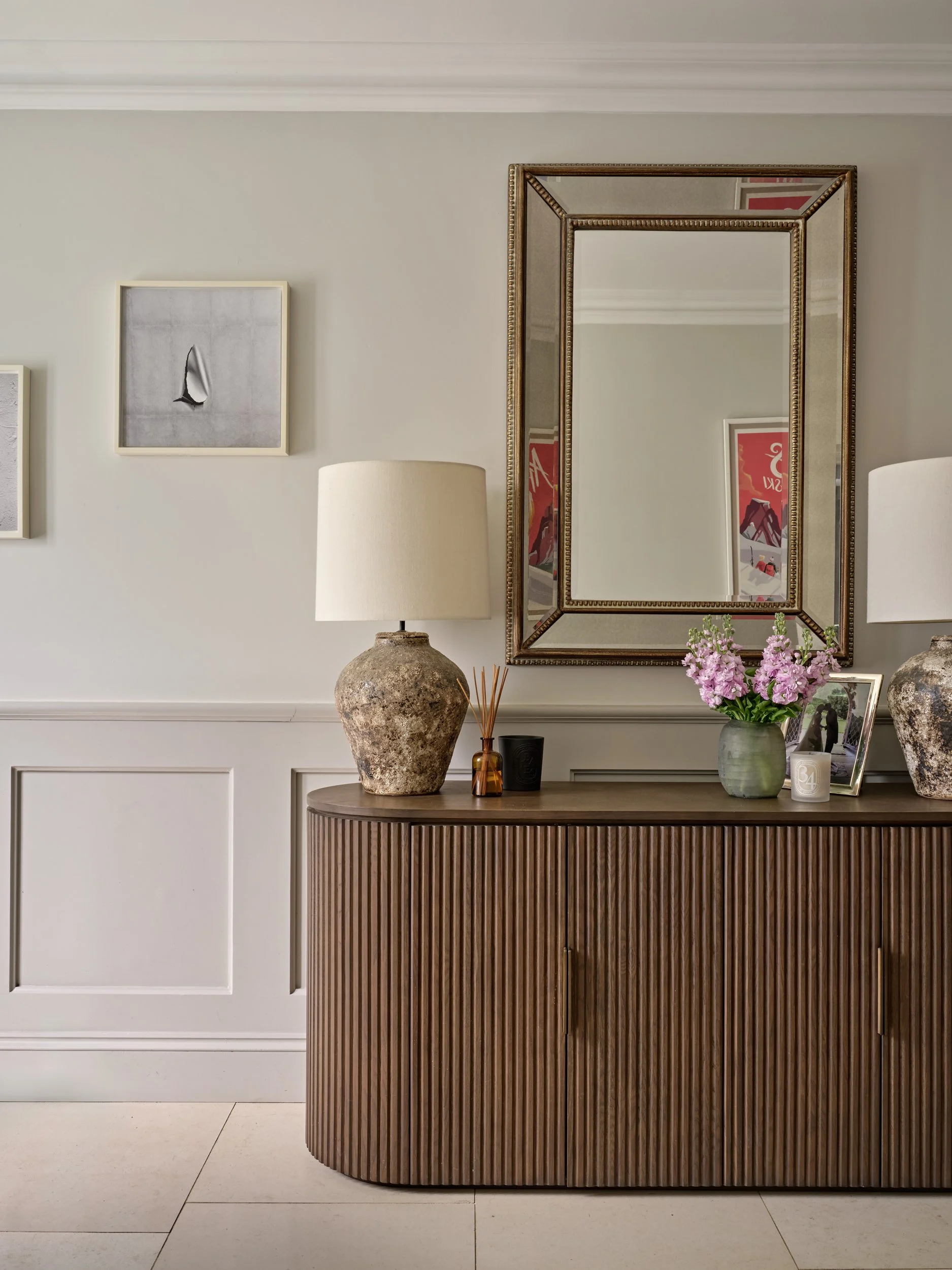 A room corner with a wooden cabinet, decorative objects, a mirror, a framed picture, and a flower vase on the cabinet surface.