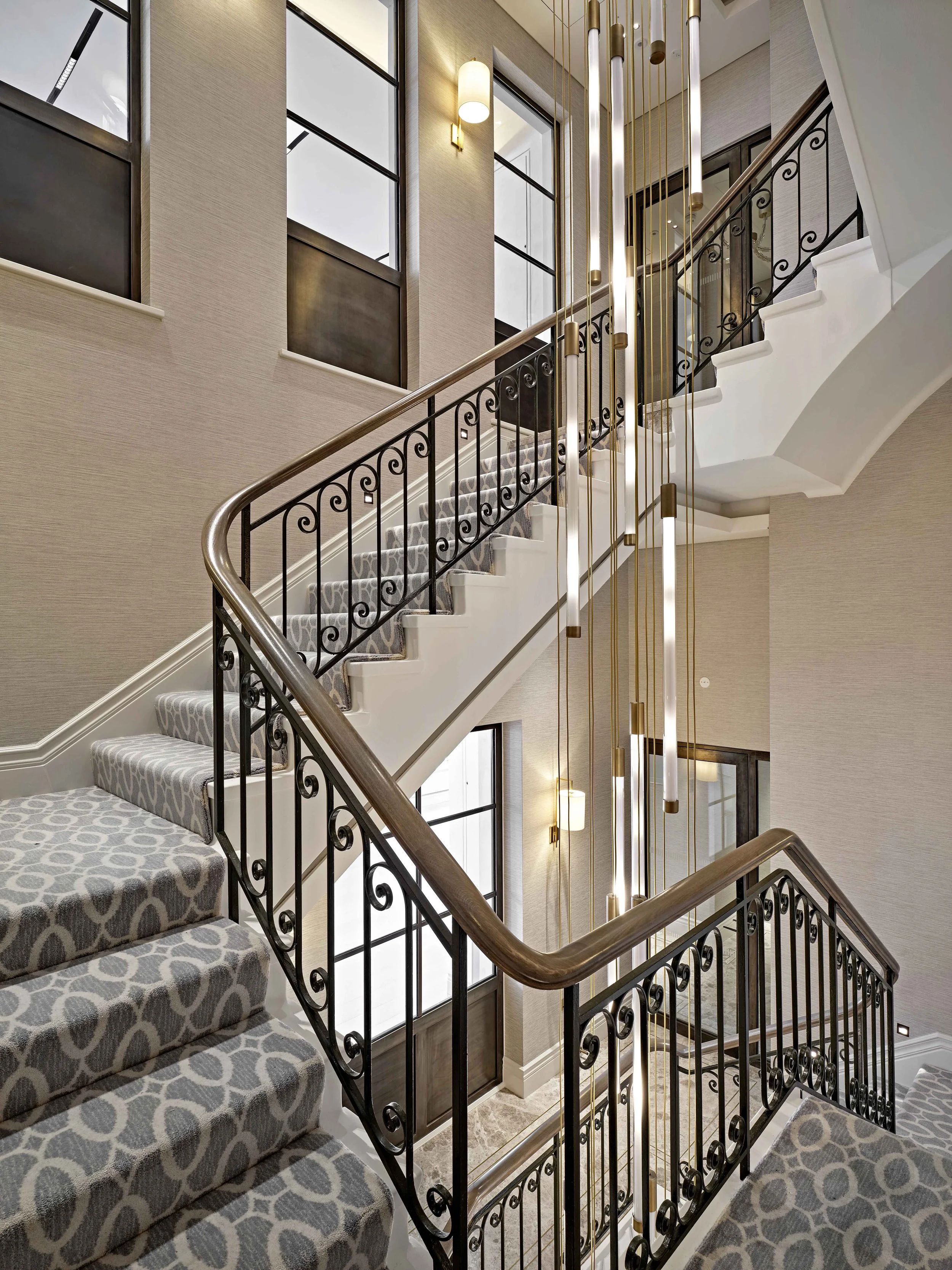 Interior view of a staircase with patterned carpet, black wrought iron railings, and large windows. Modern light fixtures hang from the ceiling, illuminating the space.