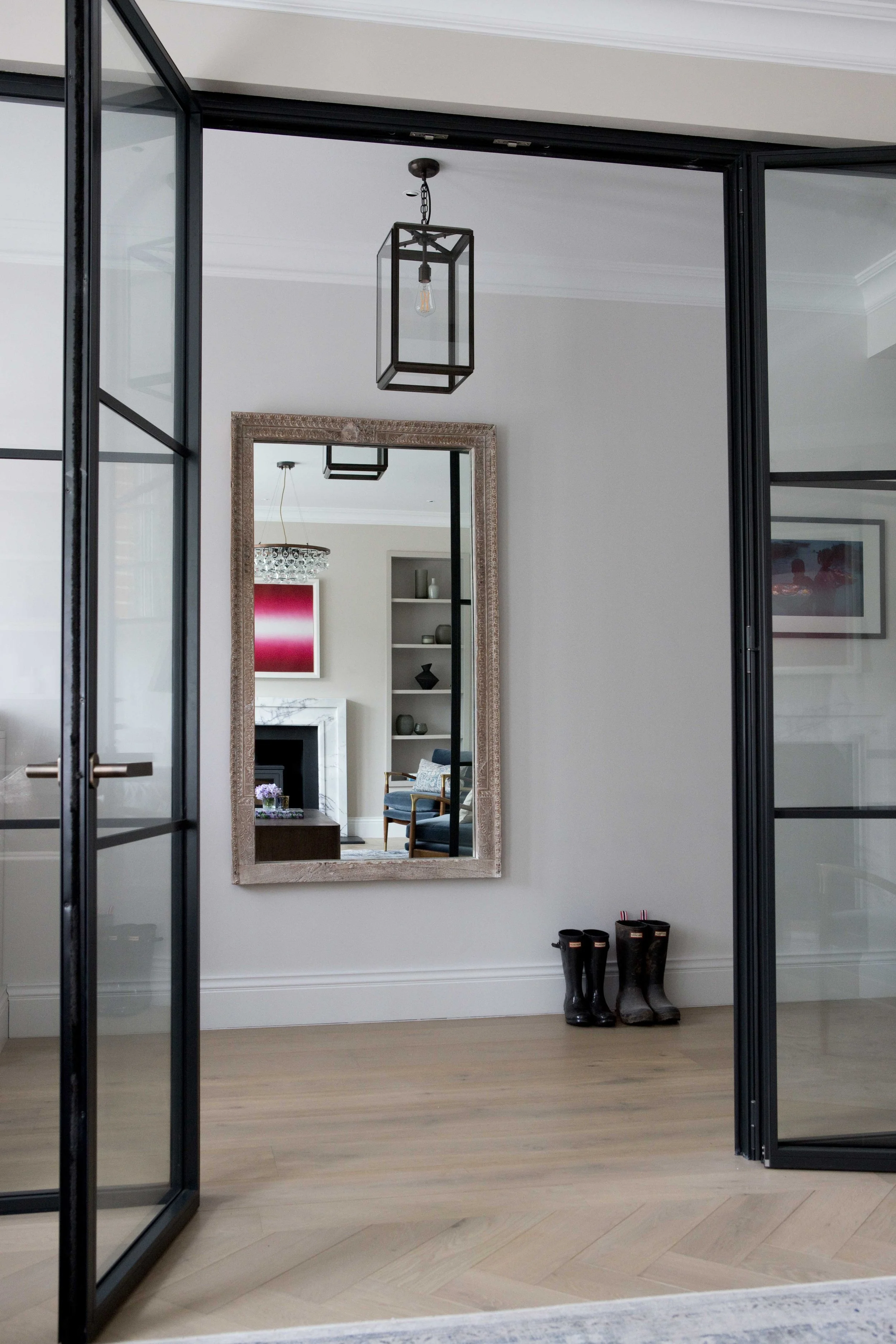 A mirror on a wall reflecting a living room with a fireplace, bookshelf, and abstract artwork. Shoes and boots are on the floor beneath the mirror. Interior space with white walls and light hardwood flooring.