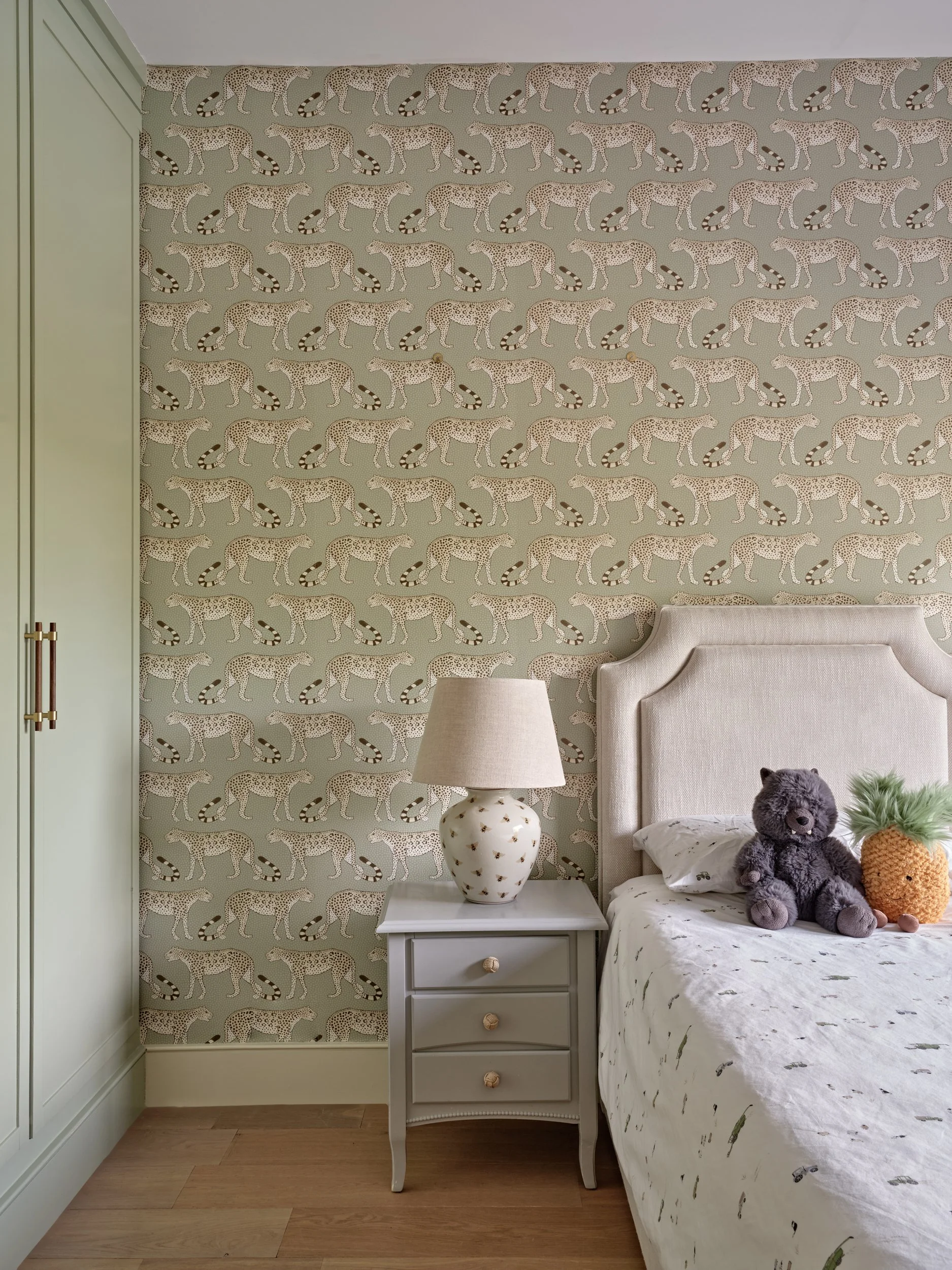 A bedroom corner with wallpaper featuring cheetahs and palm trees, a green wardrobe, a white bedside table with a starry lamp, a bed with a white bedspread, and stuffed animals including a gray teddy bear and a pineapple-shaped pillow.