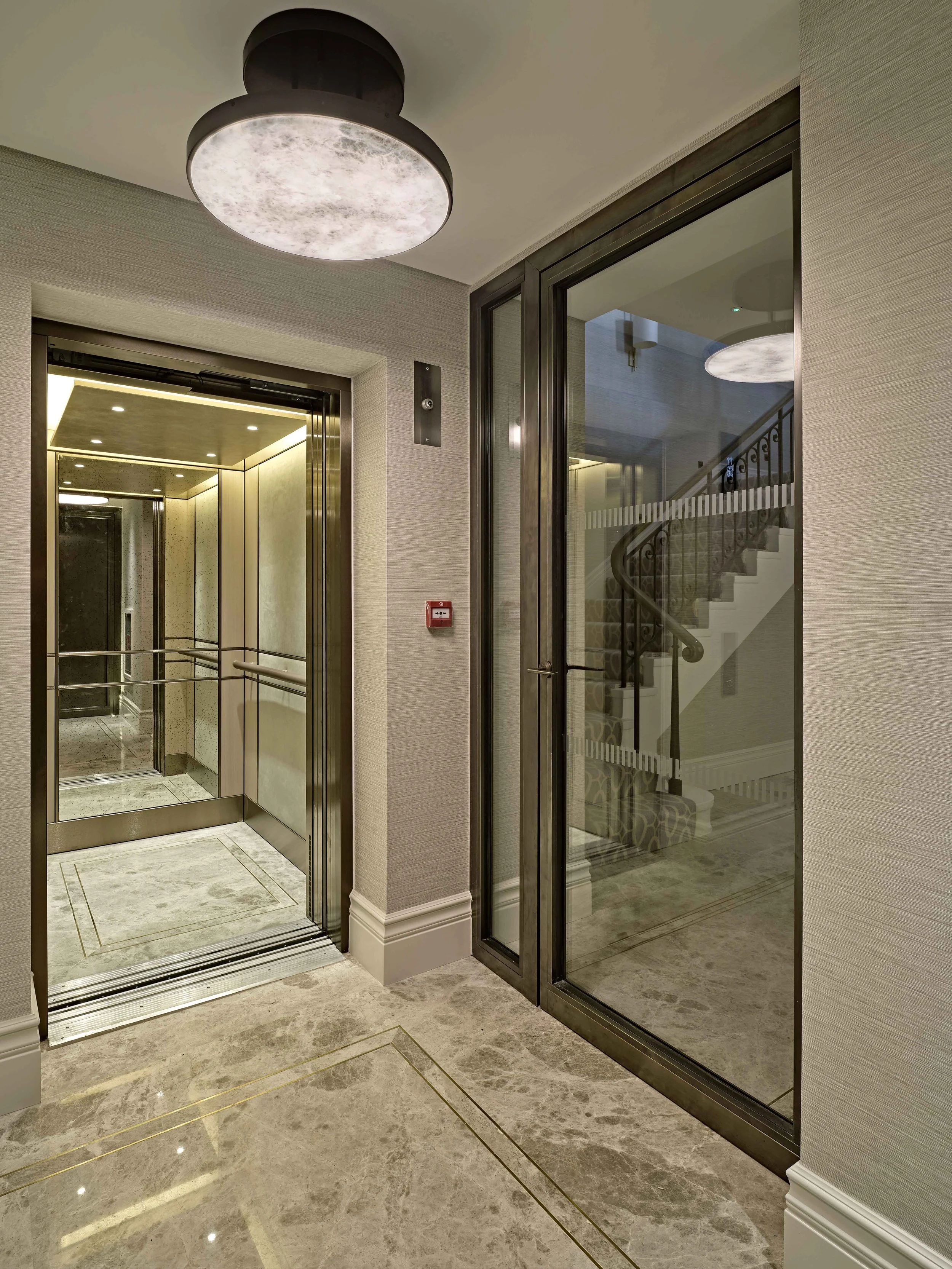 Interior of an elevator lobby showing an open elevator with beige padded walls, a reflective floor, and a corner glass door revealing a staircase inside.