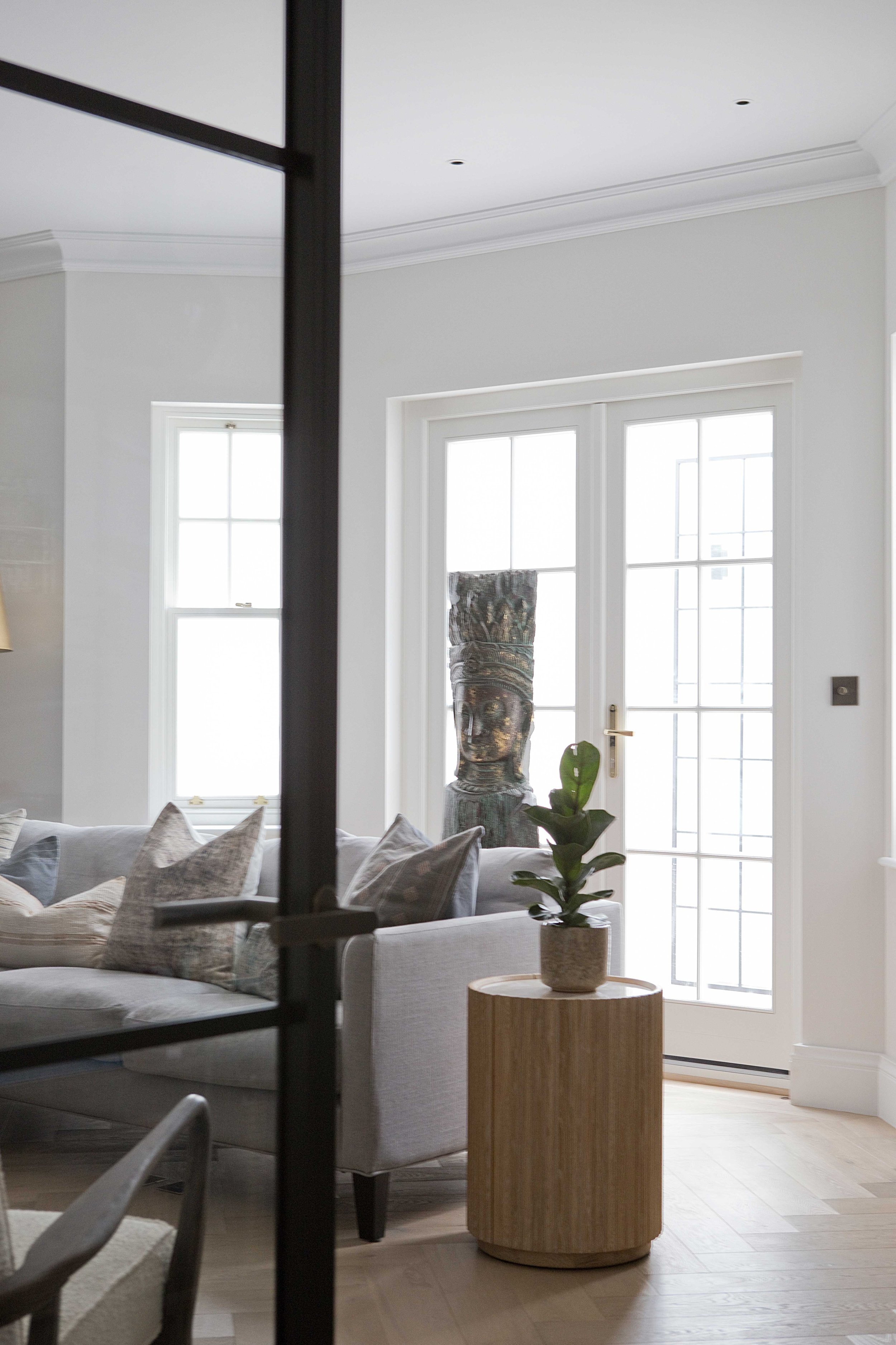 Living room with a white sofa, beige pillows, potted plant on a round wooden side table, and large windows with white frames and glass doors.