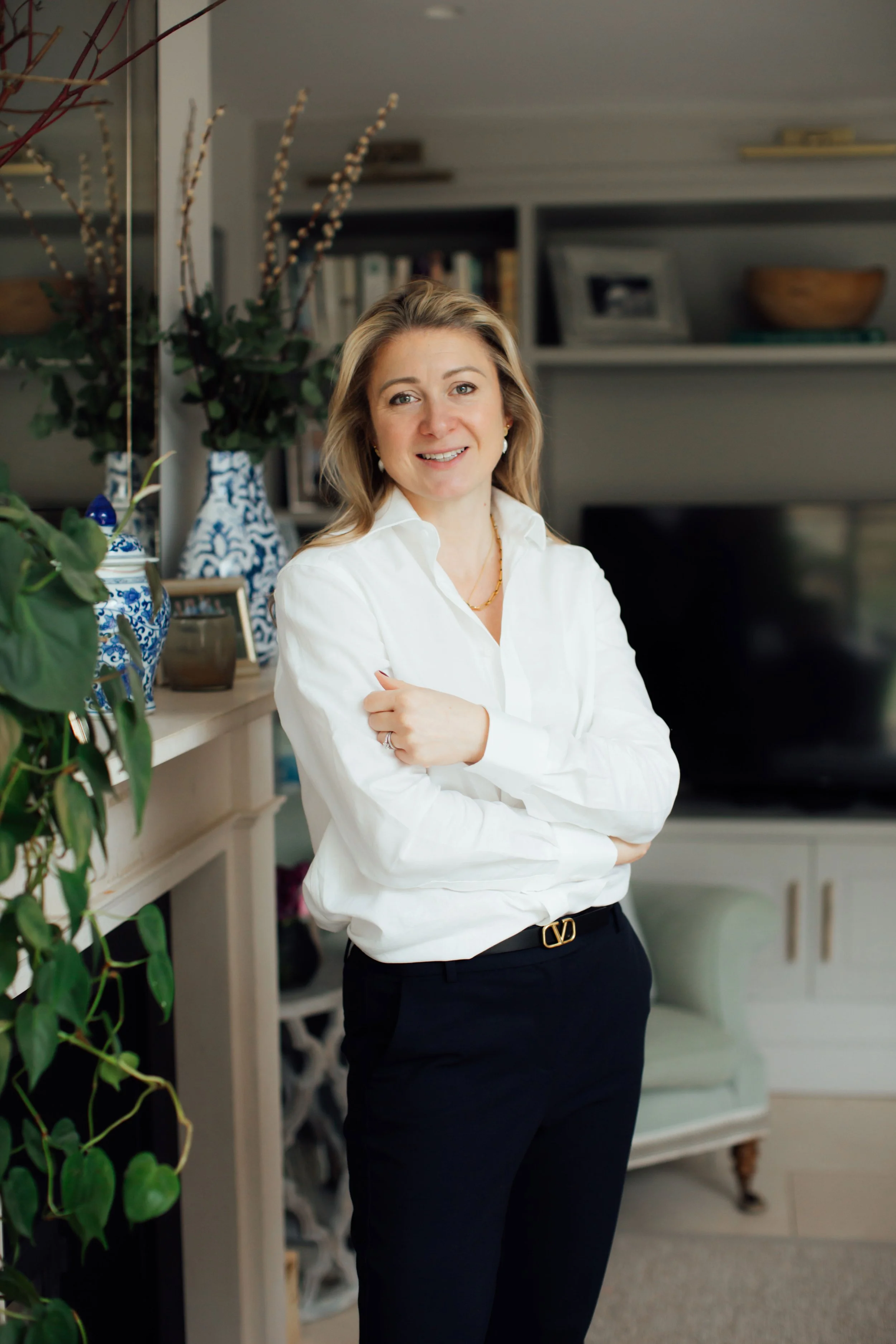 A woman with light brown hair, wearing a white blouse and black pants, standing in a room with bookshelves and decorative items, smiling at the camera.