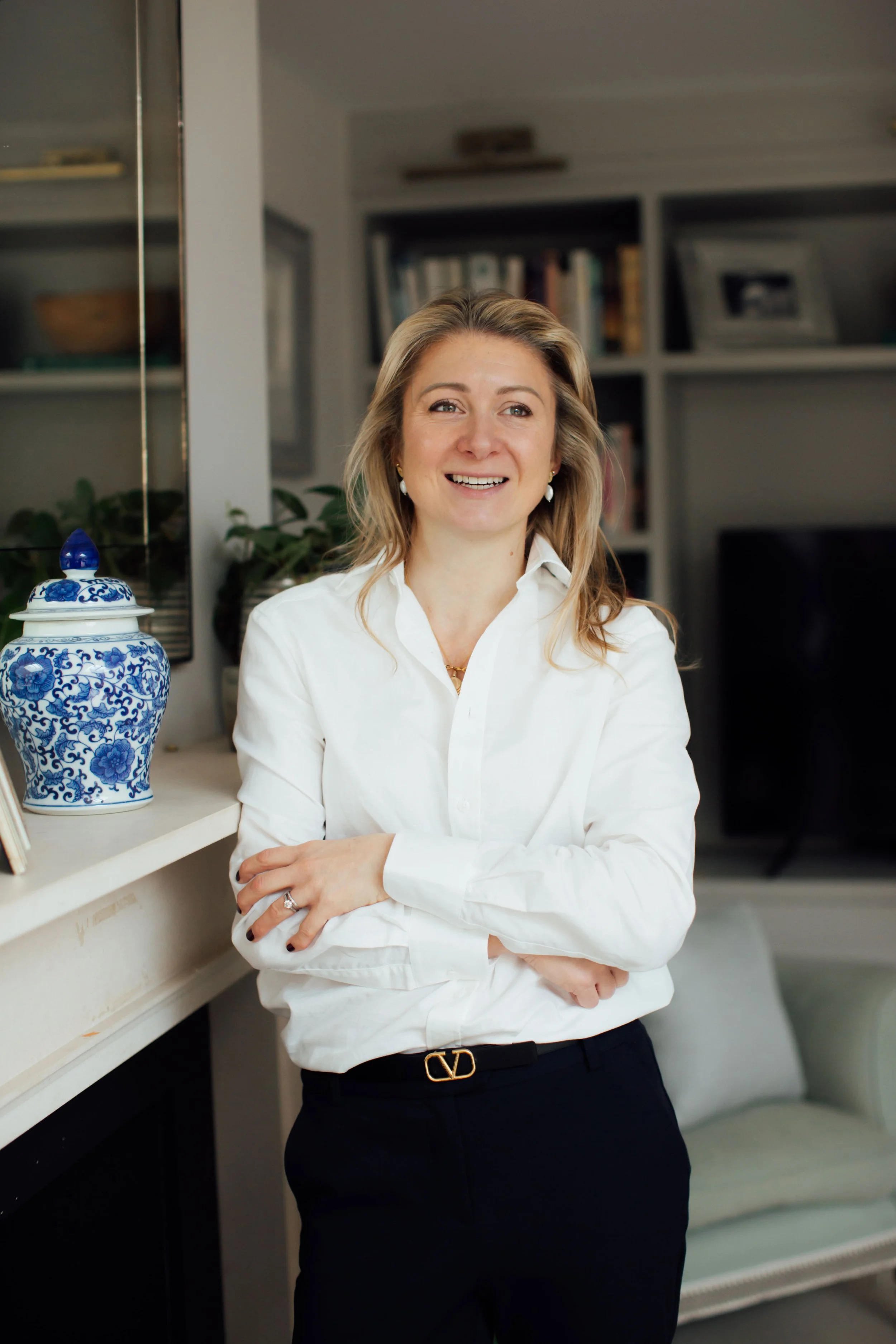 A woman with blonde hair wearing a white shirt and black pants standing indoors with crossed arms, smiling in front of a background with bookshelves, a mirror, and a blue and white porcelain vase.