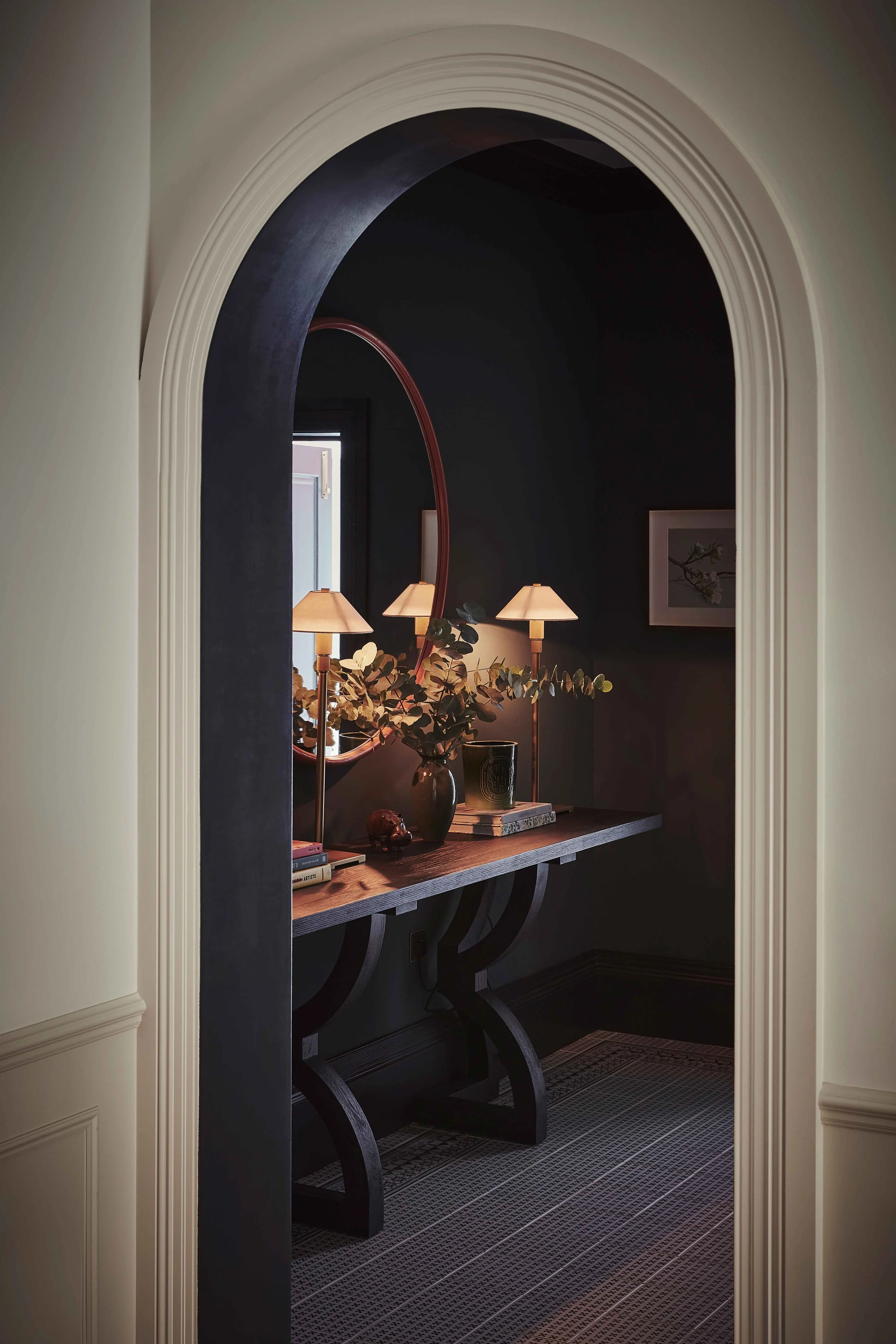 A hallway view through an arched doorway showing a dark-painted wall with a wooden console table, a round mirror, two table lamps, a framed picture, a vase with green leaves, and books on the table. The floor has a patterned dark carpet.