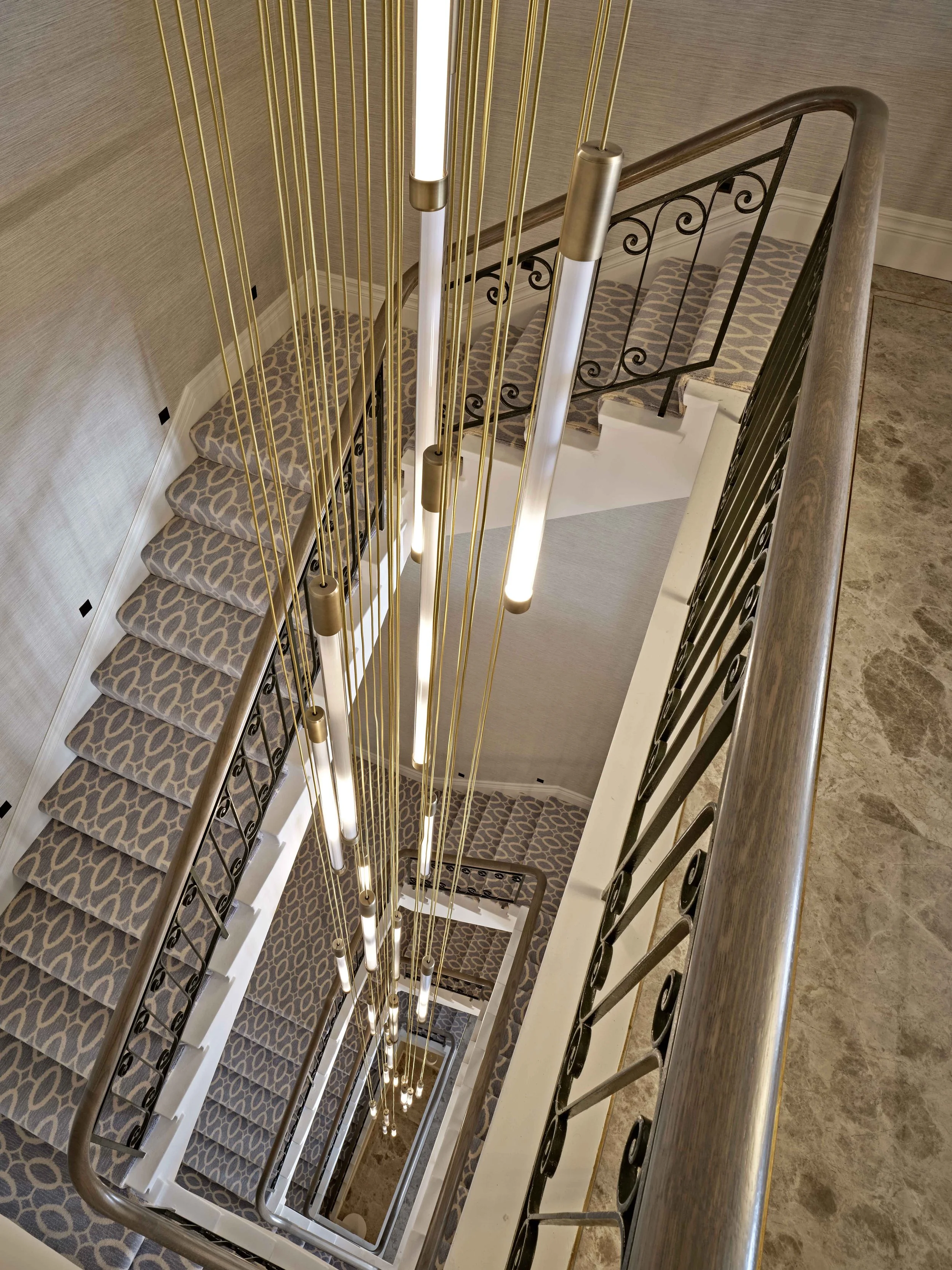 Looking down at a staircase with patterned carpet, black wrought-iron railing, and modern lighting fixtures hanging from gold-colored strings.