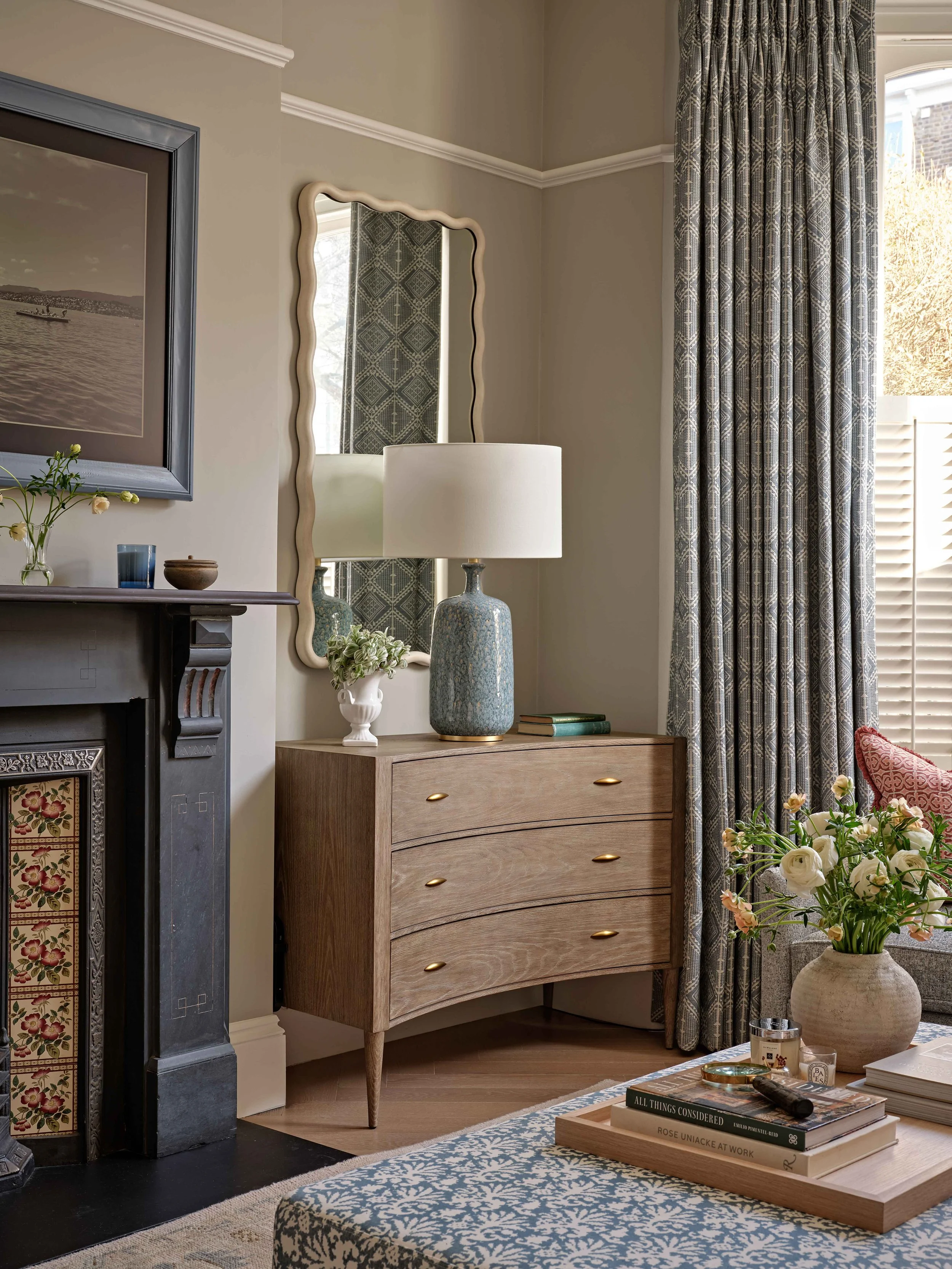 A corner of a living room with a wooden dresser, table lamp, and decorative items including a framed picture, flowers, and books, with a window and curtains on the right.
