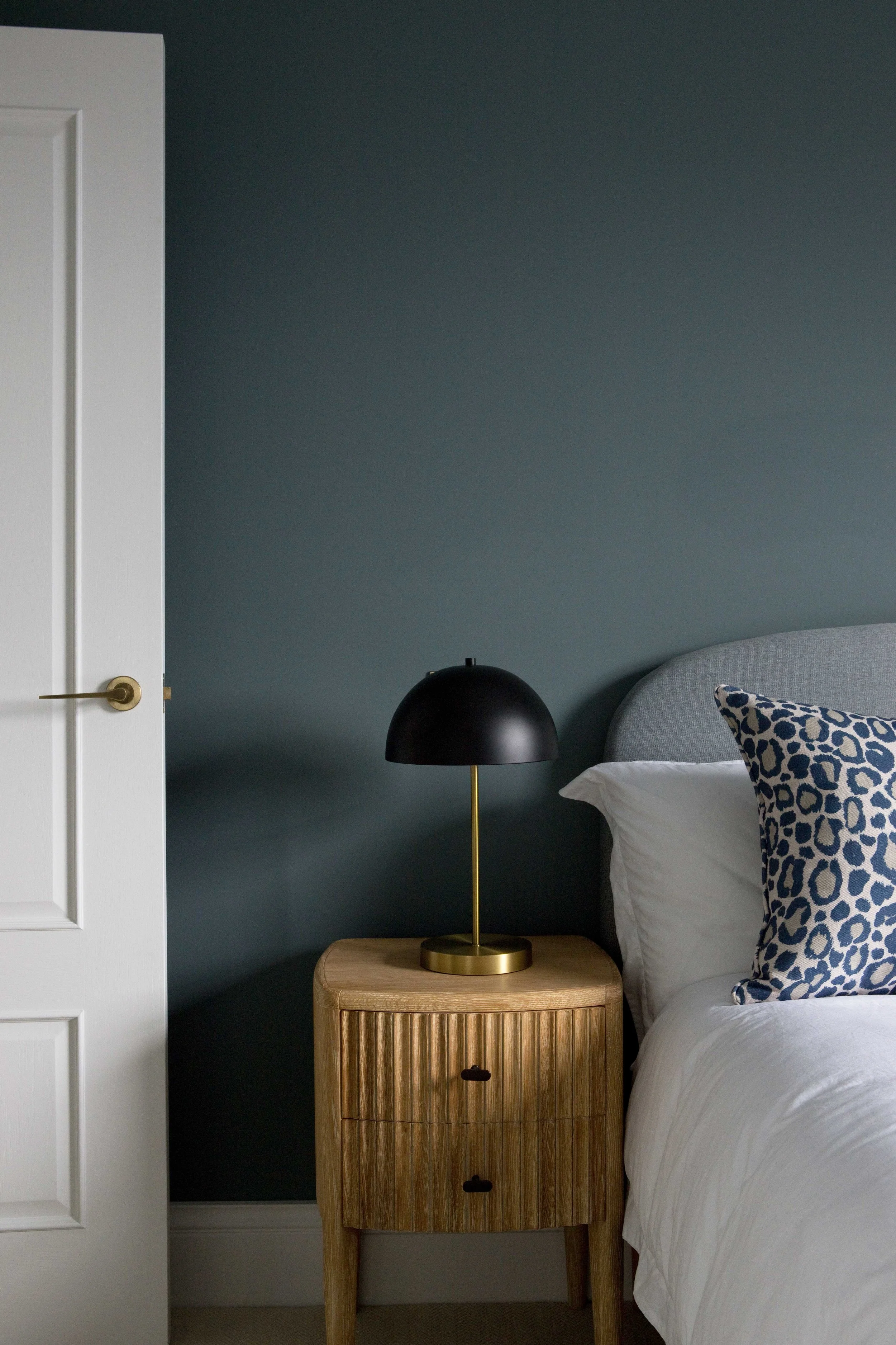 A bedroom scene featuring a wooden nightstand with a black and gold table lamp, a bed with a white duvet and a leopard print pillow, against a dark teal wall, with part of a white door visible on the left side.