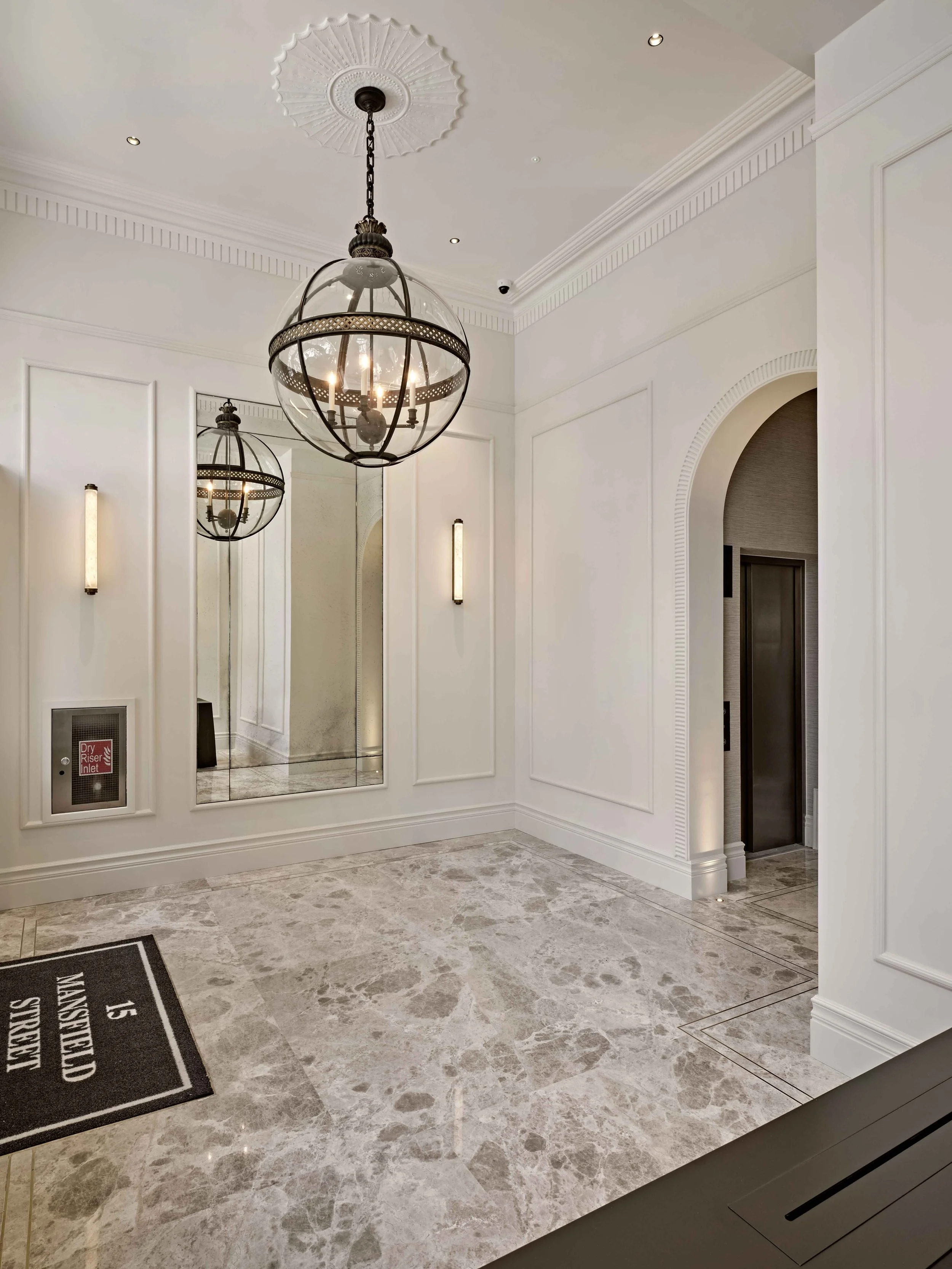 Modern interior space with white paneled walls, a large round glass pendant light fixture, wall-mounted lights, a full-length mirror, elevator, and marble flooring.