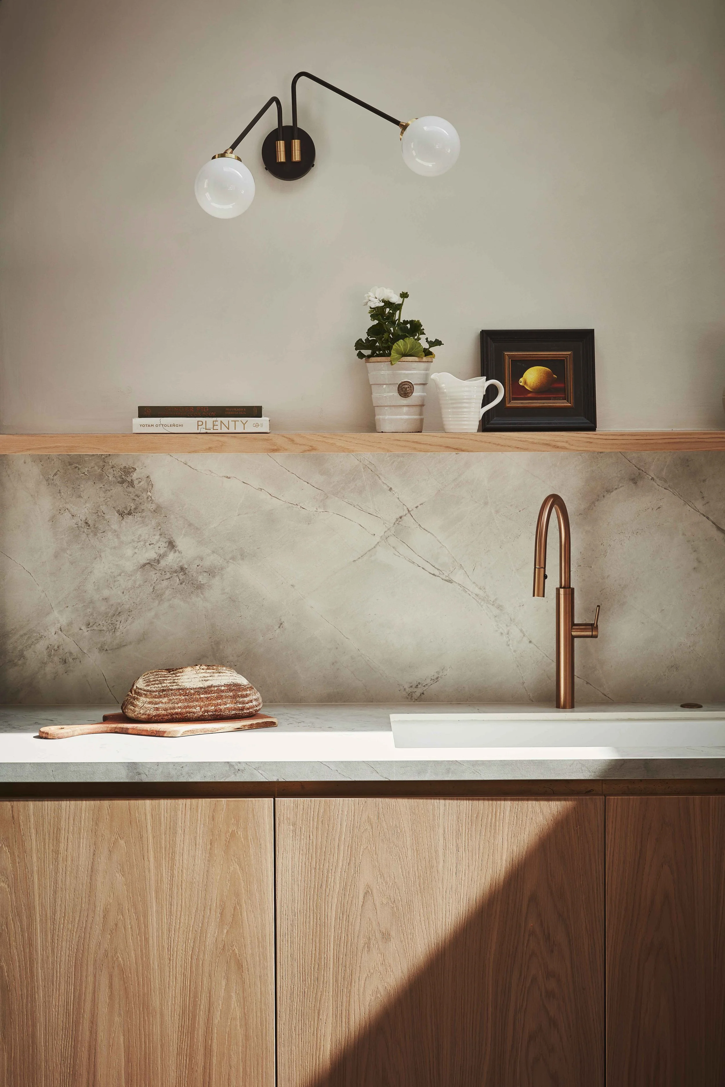 A kitchen countertop with a light wooden cabinet, a marble backsplash, a white sink with a rose gold faucet, a small stack of two stones on a wooden tray, a potted plant, a white pitcher, a framed picture of a lemon, and a modern black and brass wall