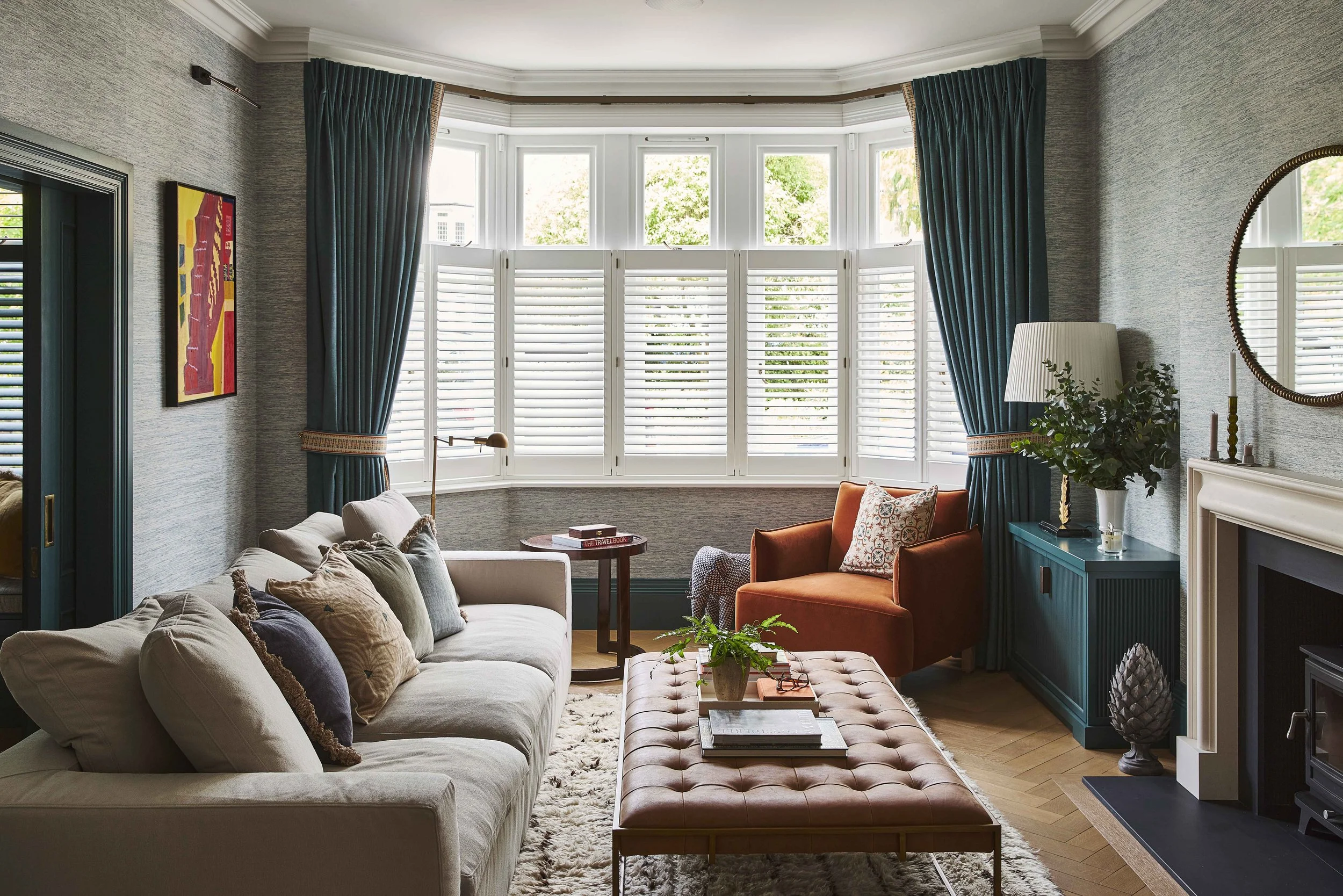 A cozy living room with beige sofa, orange armchair, tufted ottoman, and large bay window with white shutters and teal curtains, decorated with plants, books, and artwork.