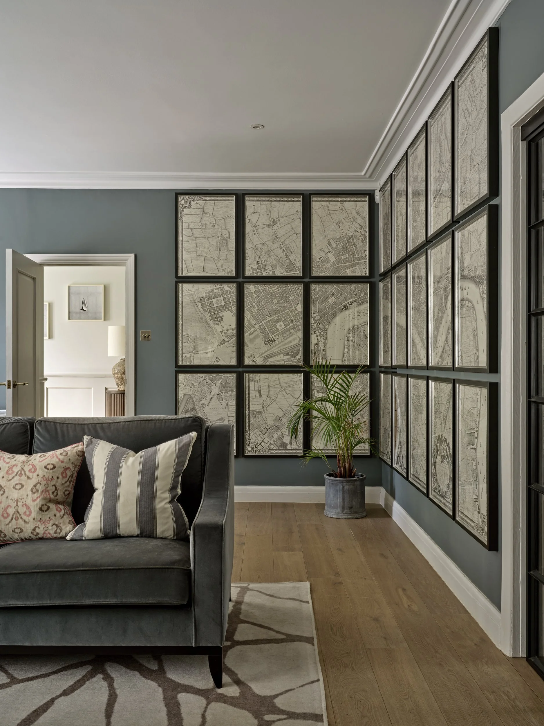 Living room with a gray sofa and patterned pillows, a rug, and a wall decorated with framed maps or city prints. There is a doorway leading to another room with a side table and a vase, and a potted plant near the maps.