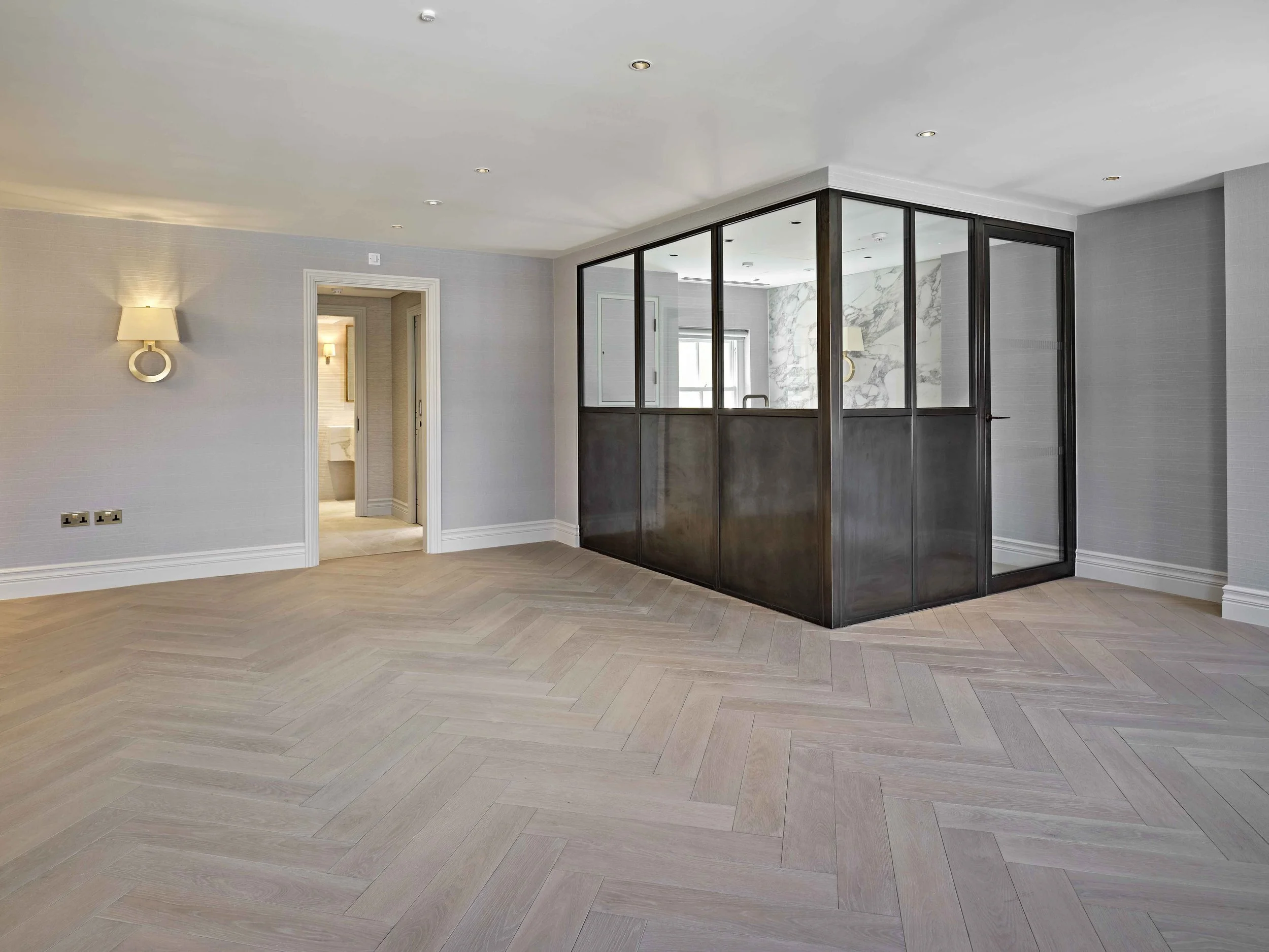 Modern living room with light-colored wood herringbone flooring, gray walls, wall sconces, and a glass partition with dark framing separating another room with marble wall accents.