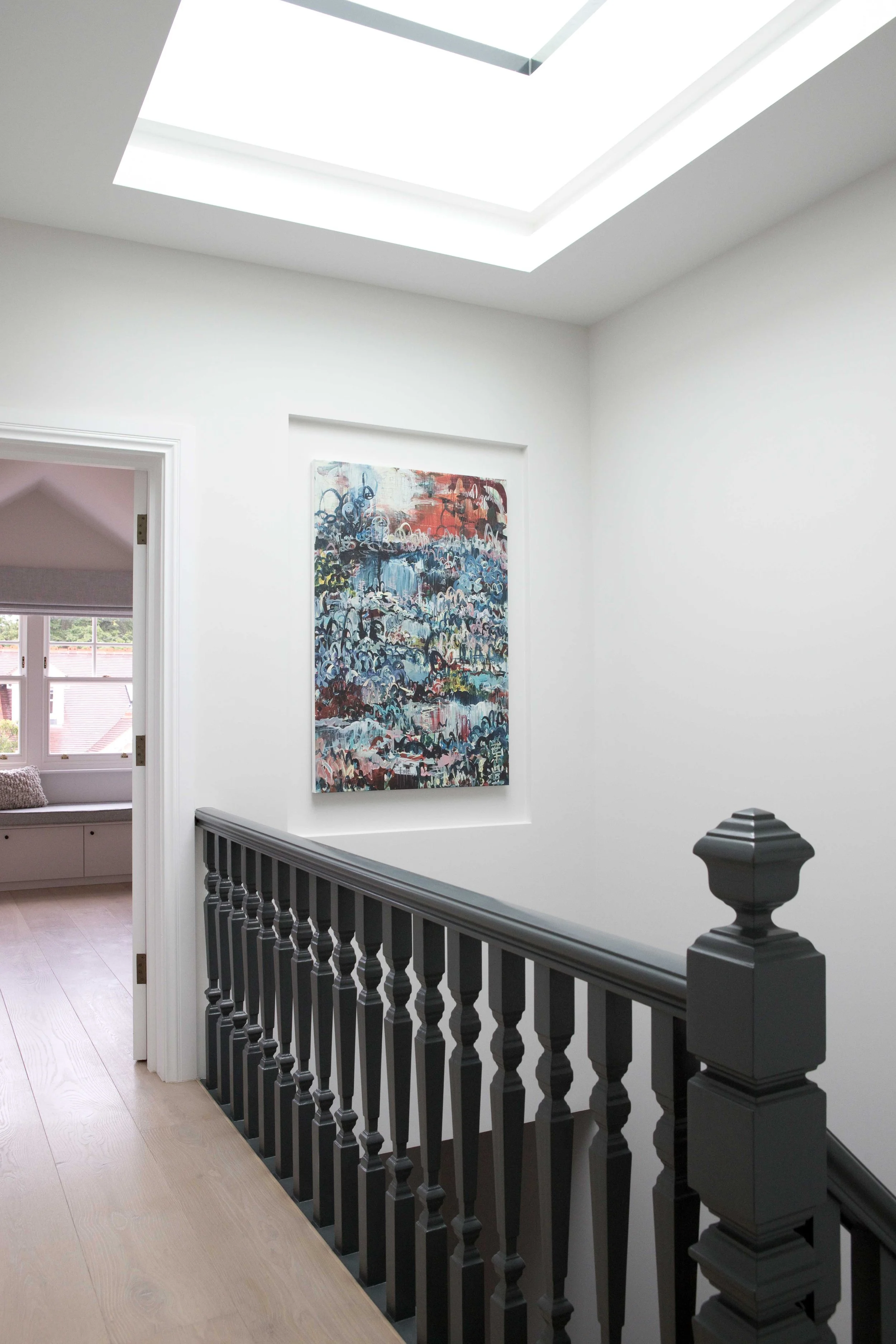 Interior view of a modern home's staircase landing with white walls, a dark gray railing, a colorful abstract painting on the wall, and a skylight ceiling.