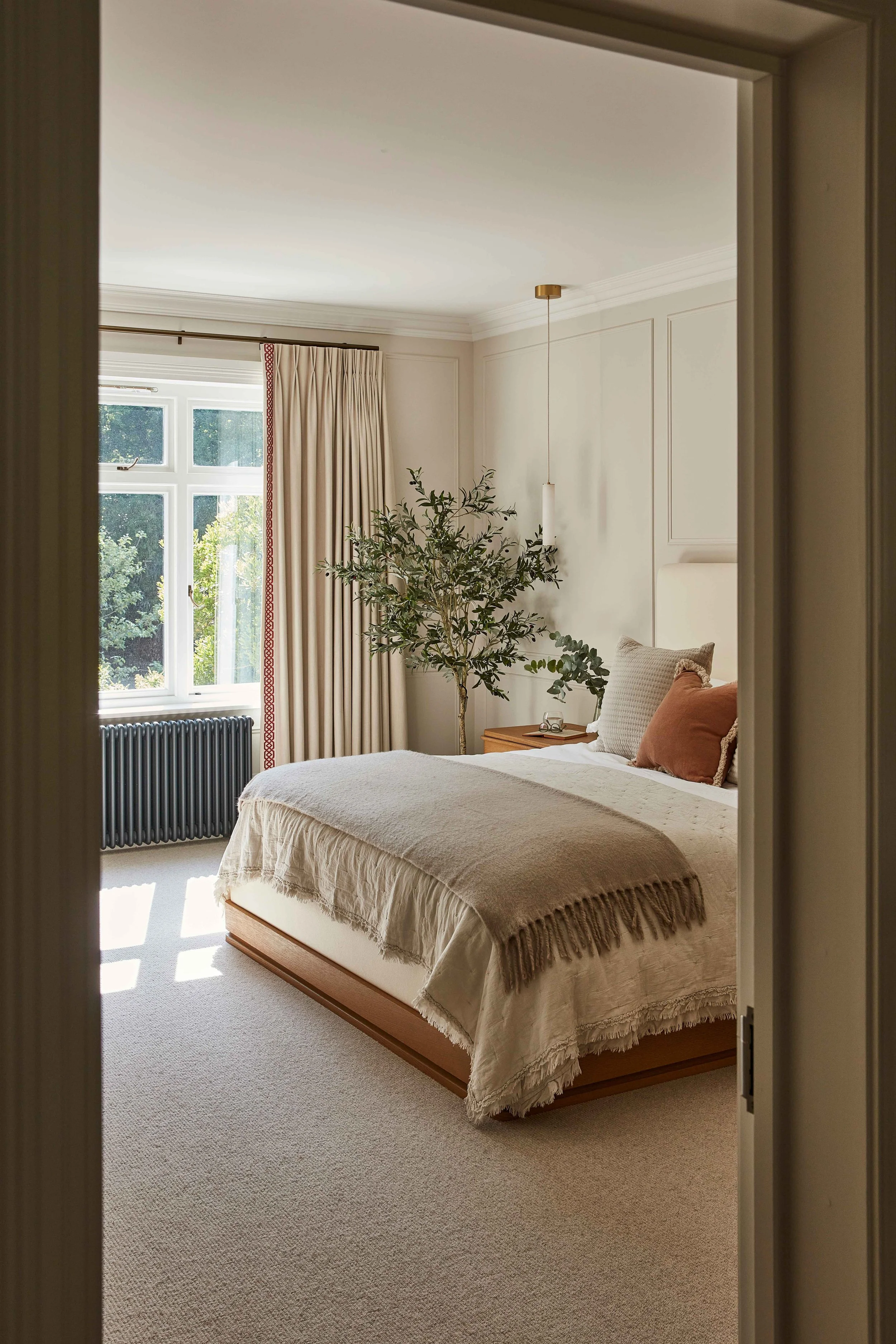 A cozy bedroom with a large bed covered in beige textured bedding, set against a wall adorned with white paneling. There are multiple pillows on the bed, including a large cream pillow and a smaller rust-colored one. A wooden nightstand beside the be