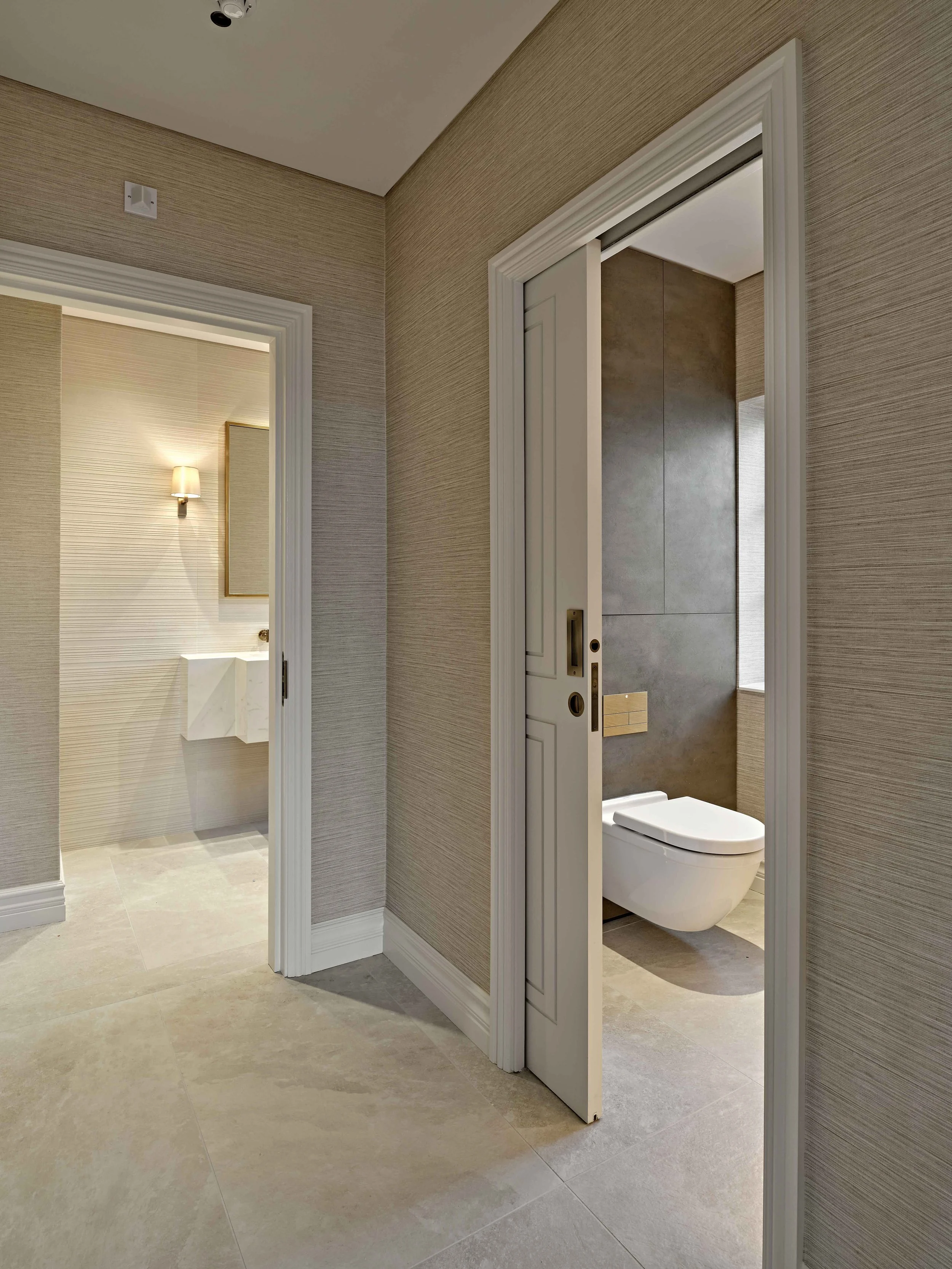 Interior view of a modern bathroom with a sliding door partially open, revealing a toilet and a beige textured wall. The space is decorated with neutral tones, and there is a wall-mounted light fixture and a framed mirror visible through the door on 