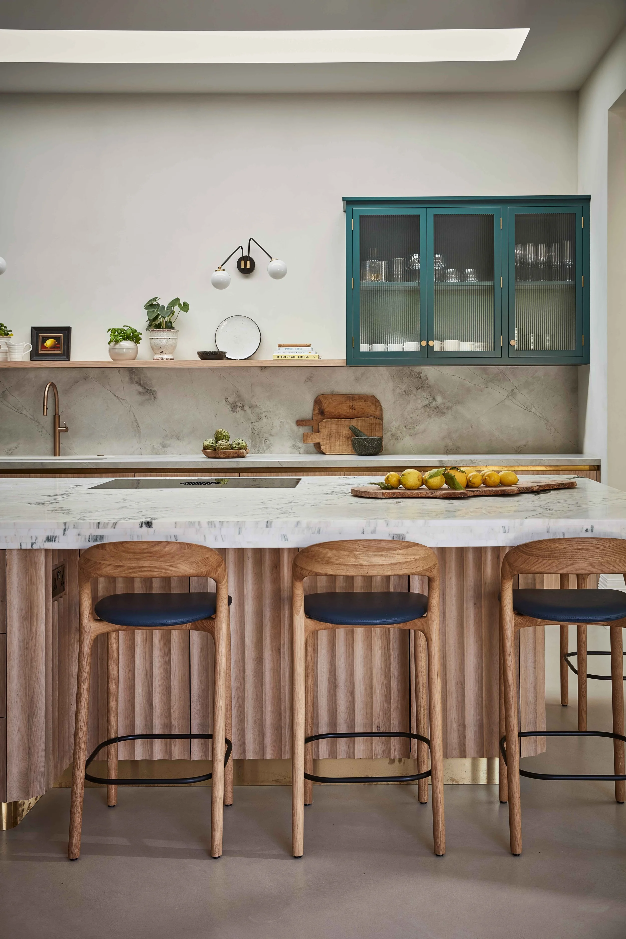Modern kitchen with white marble countertop, wooden bar stools, a light blue cabinet, potted plants, decorative plates, a small cutting board with lemons, and a wall-mounted lamp.