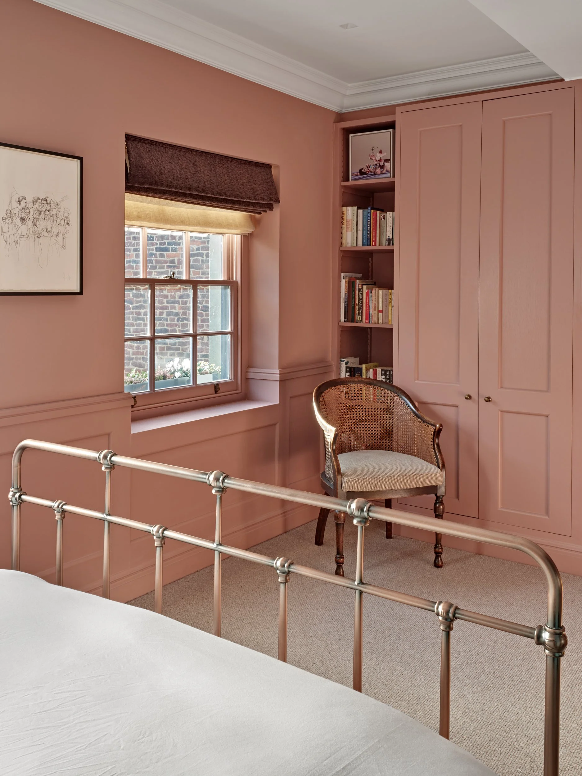A bedroom corner with pink walls, a window with a brown and beige Roman shade, built-in bookshelves filled with books, a pink cabinet, a beige armchair, and part of a metal bed frame.